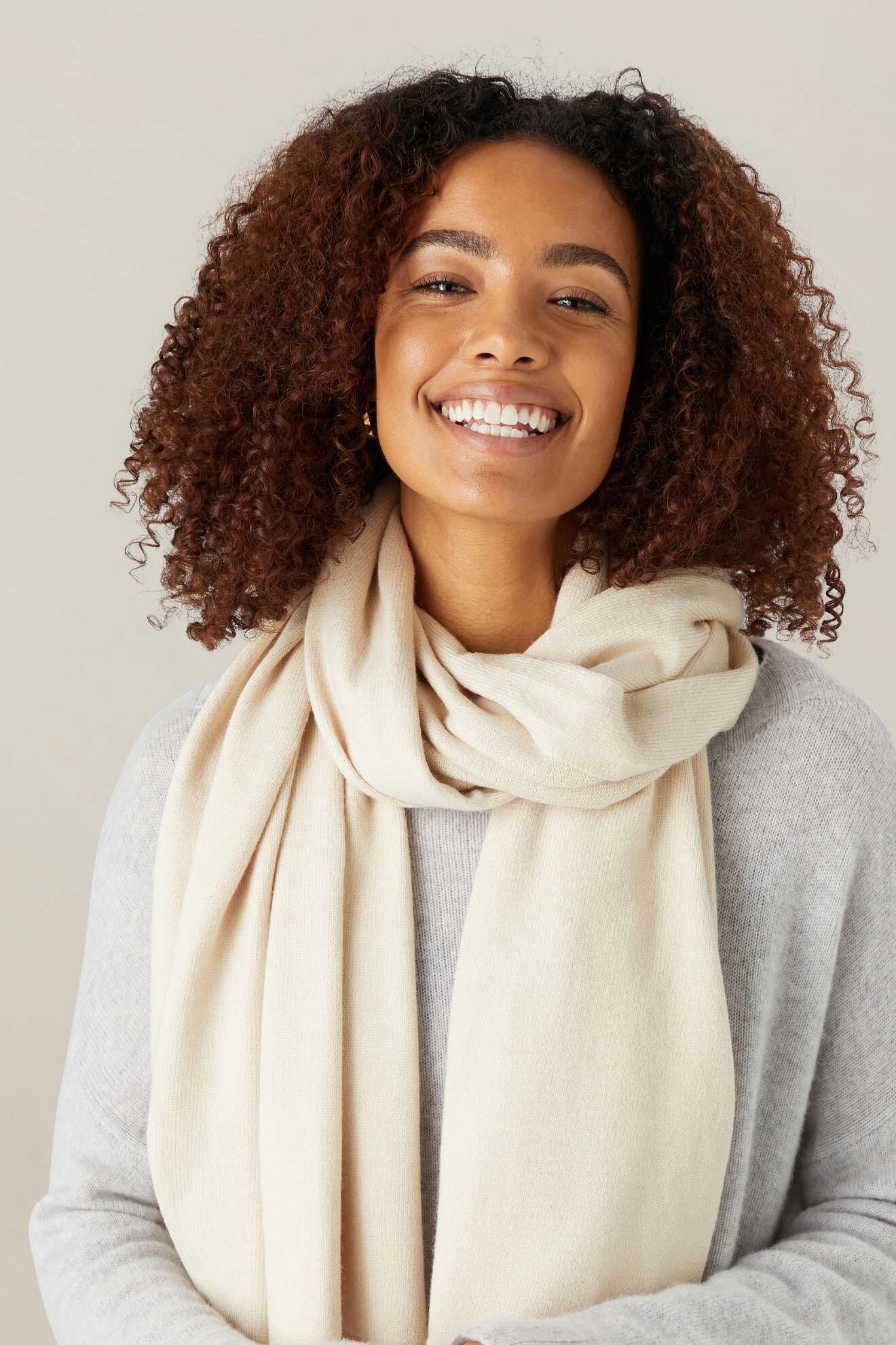 Woman with curly hair smiles at the camera, wrapped in a Knit Scarf and gray sweater—her go-to winter warmer against a neutral background.