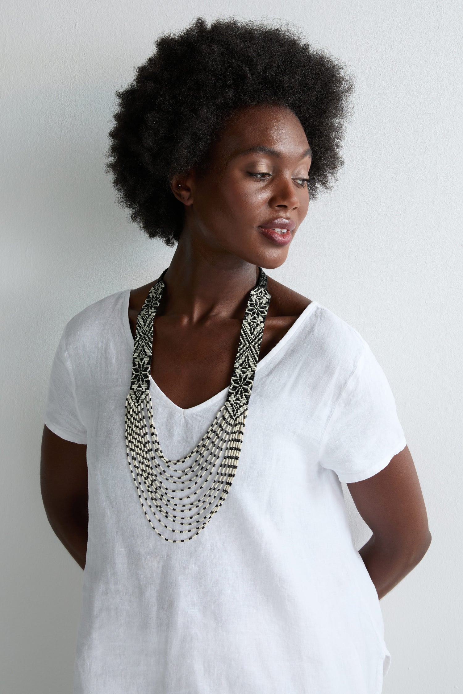 A woman with short curly hair and a white blouse showcases the Artisanal Aztec Ebony Beaded Necklace—hand-beaded panels in a bold monochrome palette—against a plain light background.
