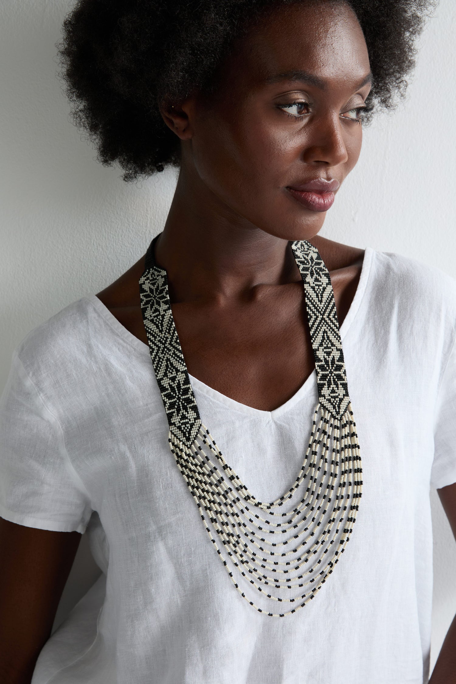 Woman in a white v-neck top wears the Artisanal Aztec Ebony Beaded Necklace with hand-beaded panels, gazing to the side against a plain background for a striking monochrome look.