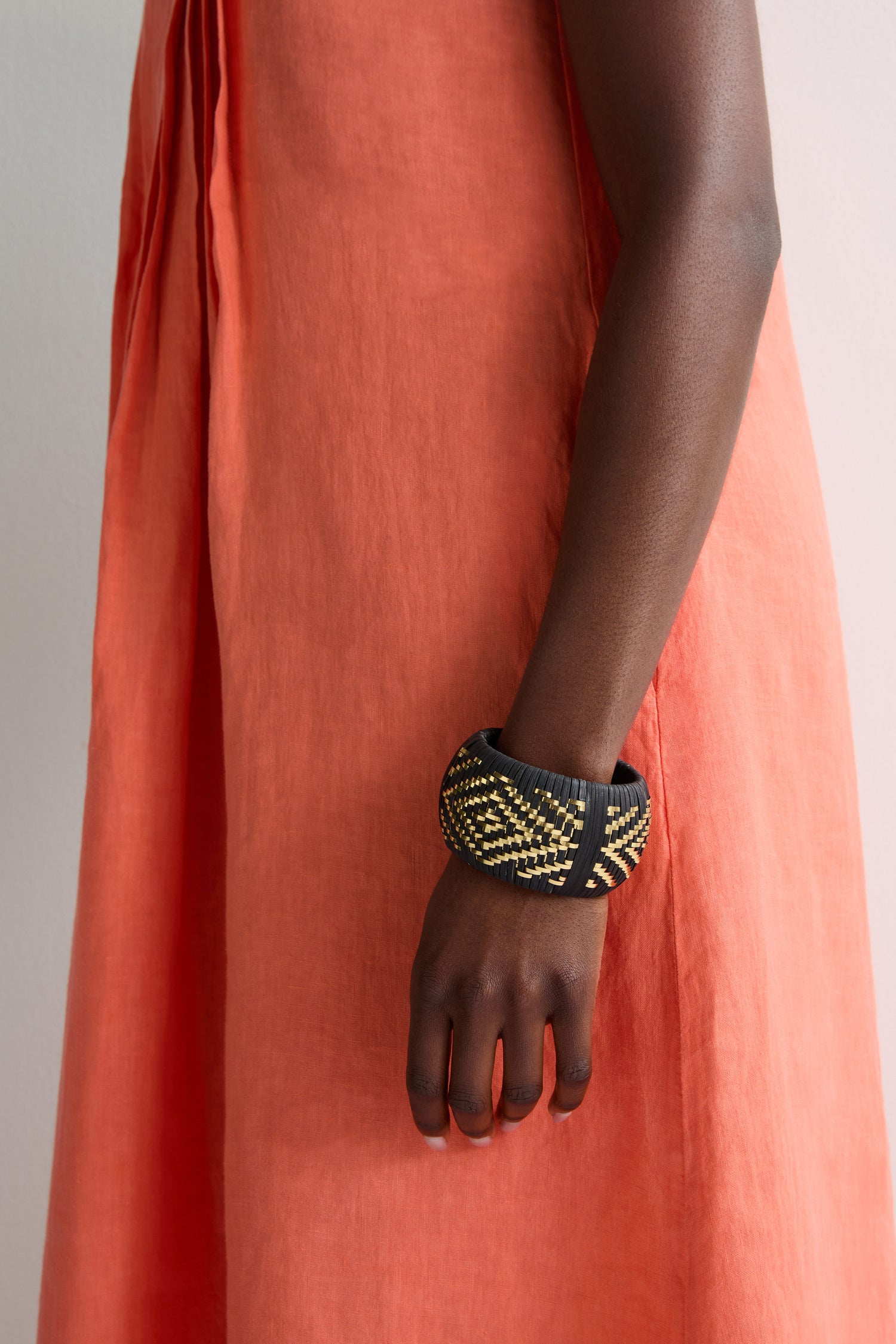 A person wearing an orange dress and the Handwoven Gold Detail Bangle with black, yellow, and diamond patterns stands against a plain background.