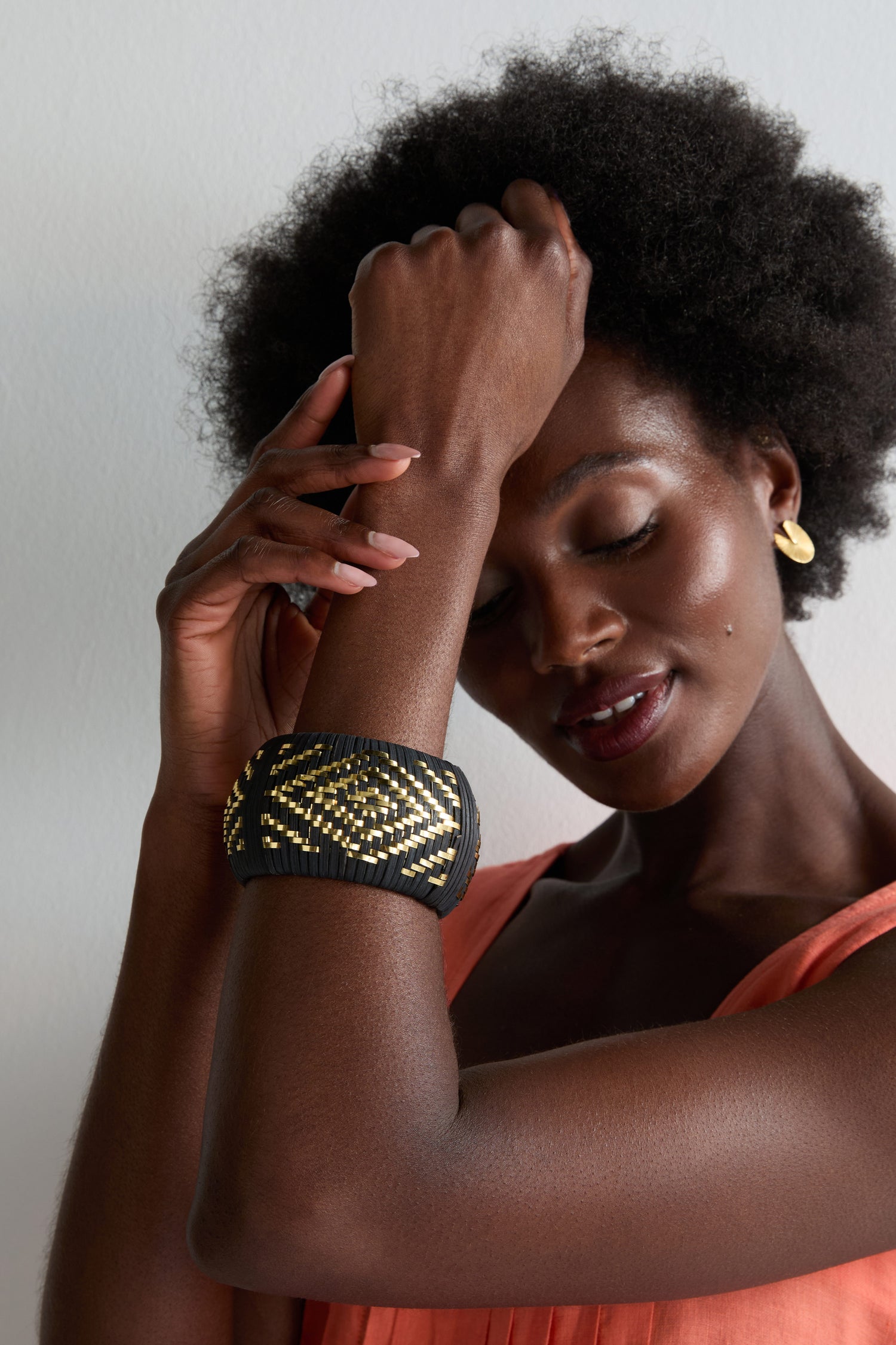 A woman with an afro hairstyle wears a sleeveless orange top, gold earrings, and the Handwoven Gold Detail Bangle—featuring black and gold diamond patterns—as she poses with one arm raised and her eyes gently closed.