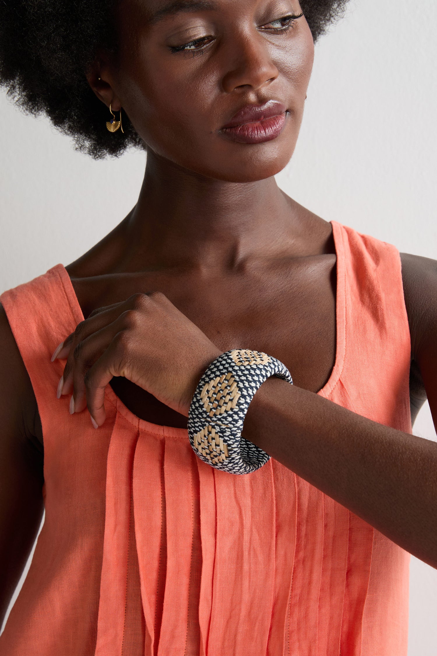 A woman wearing a sleeveless coral top looks down at her raised arm adorned with the Handwoven Rattan Detail Bangle in black and beige.