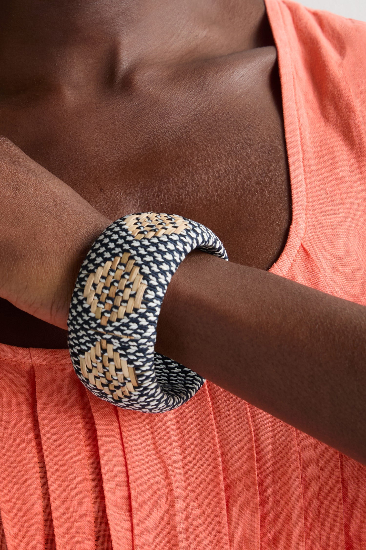 A person wearing a coral pleated top and a Handwoven Rattan Detail Bangle, a statement accessory with black, beige, and white patterns, on their wrist.