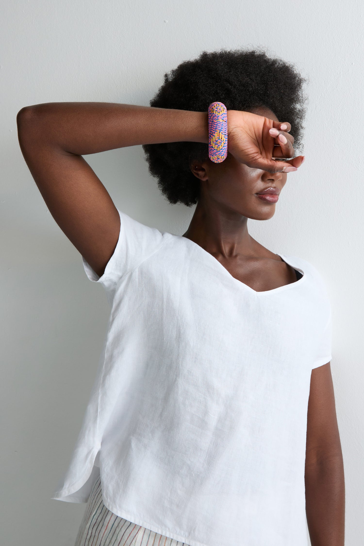 Wearing a white short-sleeved top, a person stands against a plain white background with one arm across their face, showcasing the Handwoven Fluoro Bangle in bright neon tones.