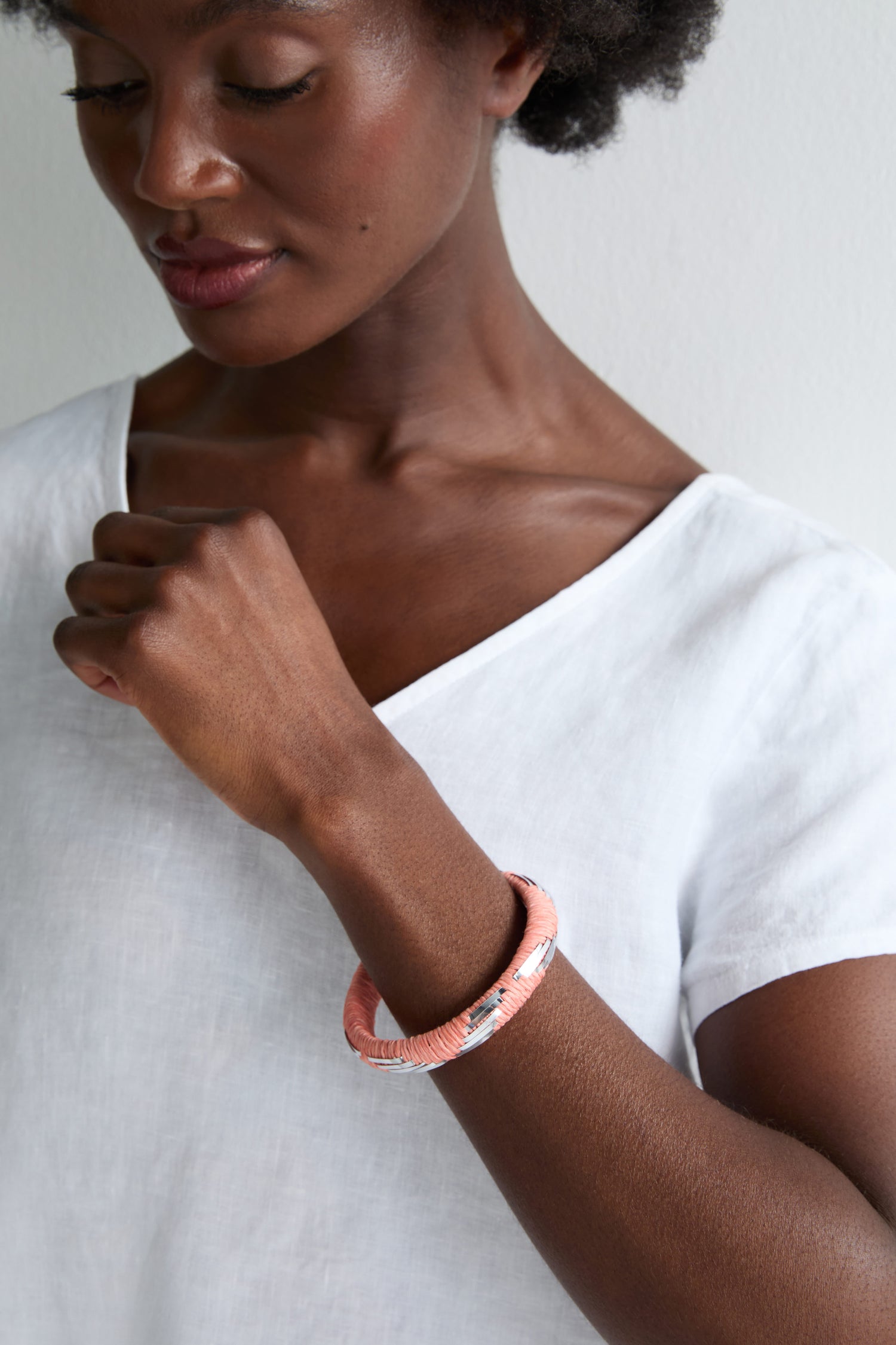 A woman in a white top looks down, showcasing the Handwoven Silver Chevron Bangle featuring silver accents and a touch of pink charm on her wrist.