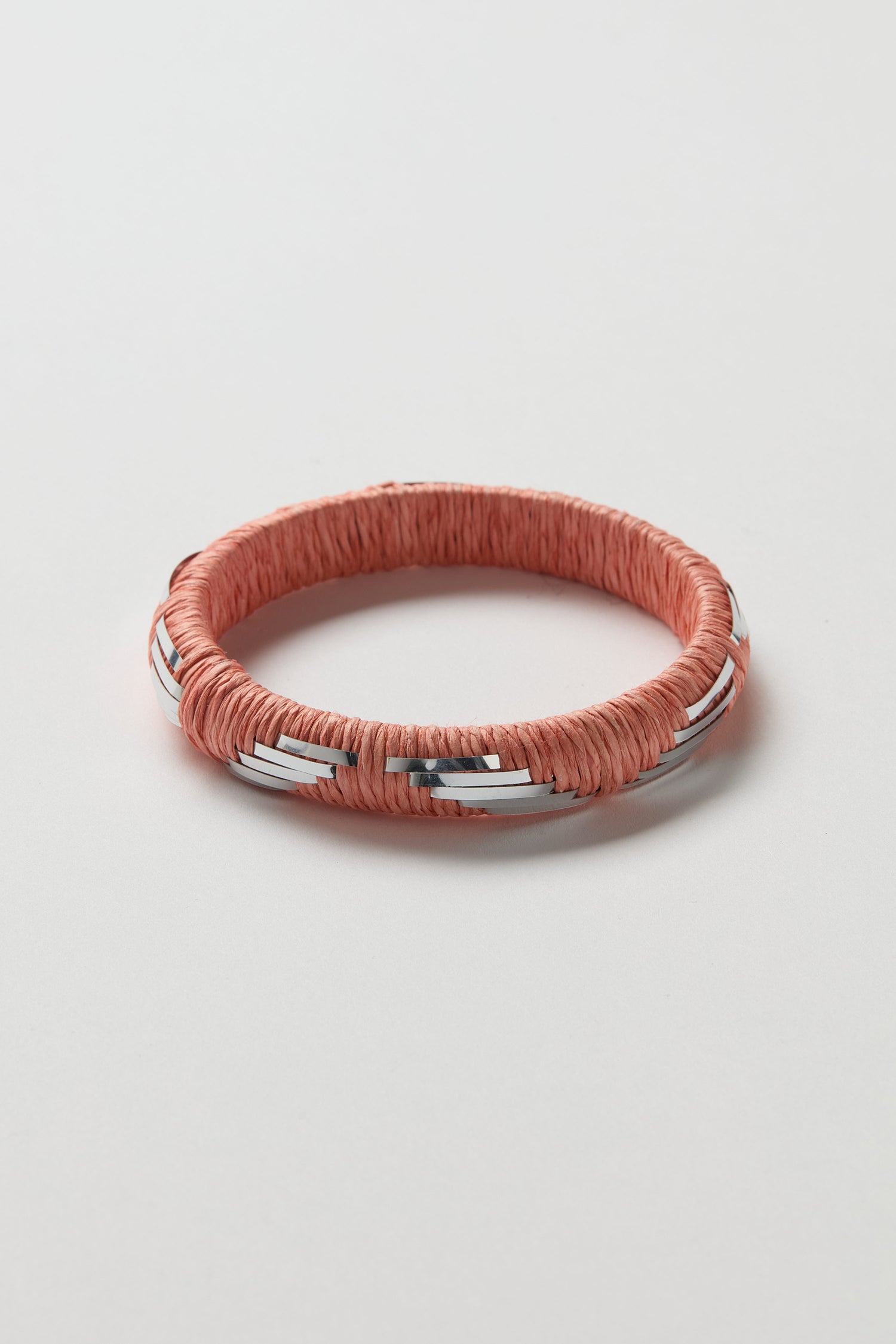 The Handwoven Silver Chevron Bangle is a round artisanal piece wrapped in pink cord with evenly spaced silver chevron accents, displayed on a white surface.