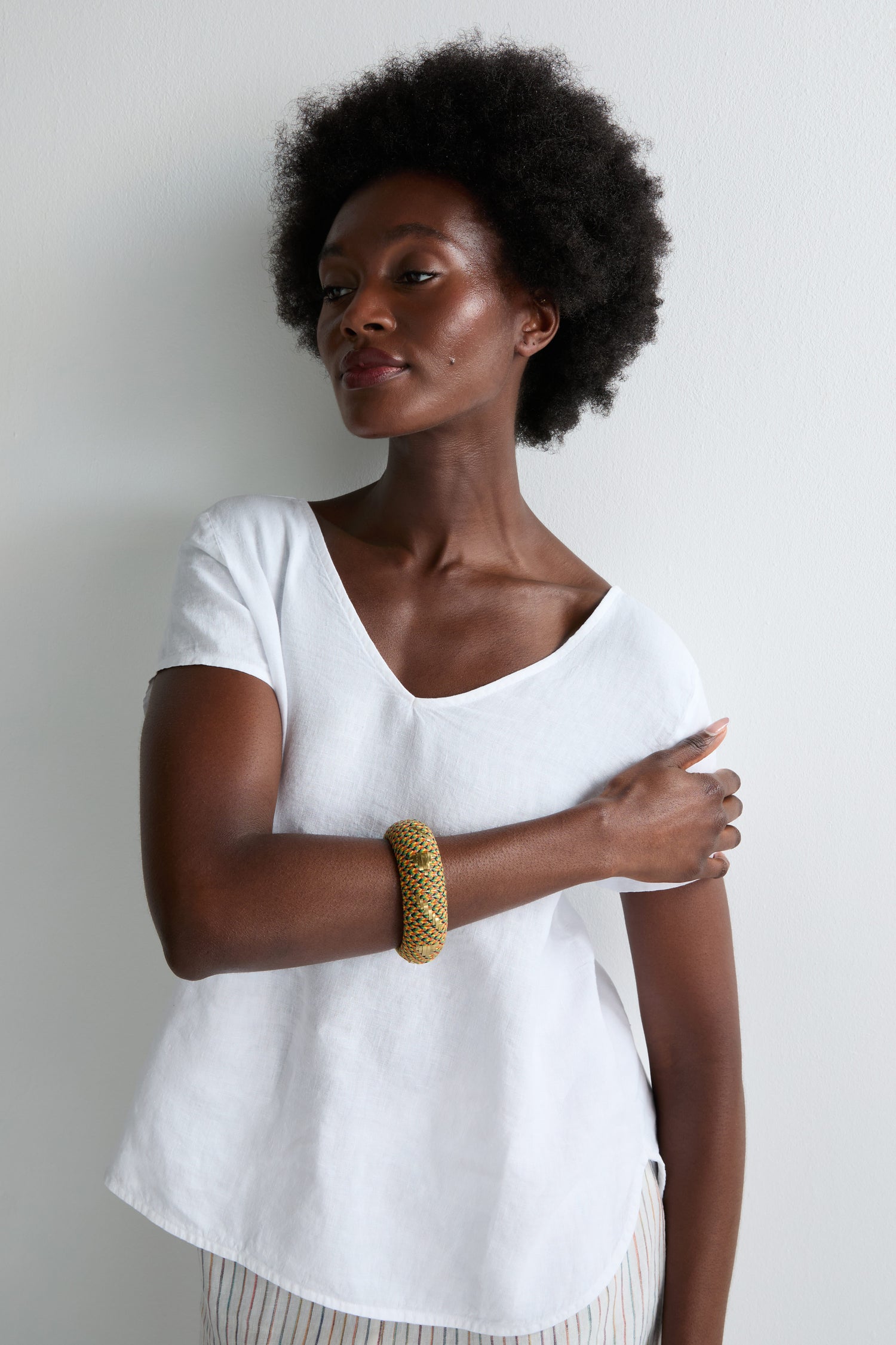 A woman with short, curly hair in a white shirt wears the Handwoven Multicoloured Bangle, known for its vibrant threads and heritage craftsmanship, as she stands against a white wall with one arm across her body.