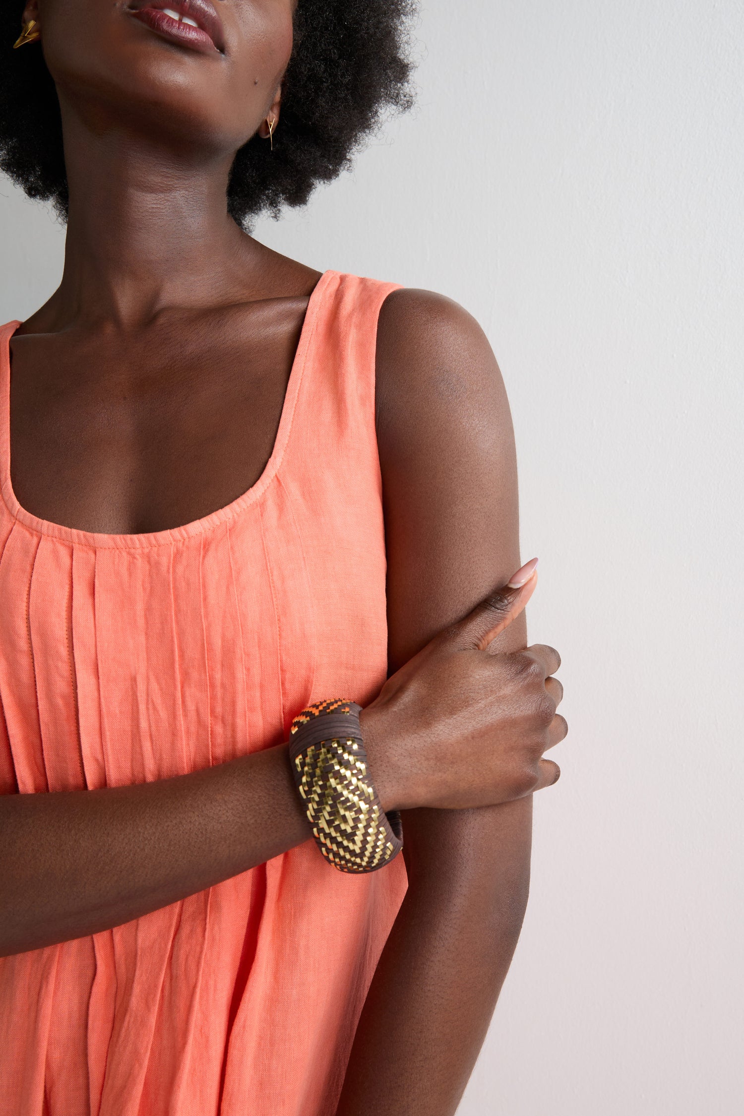 Wearing a sleeveless coral dress and the Handwoven Espresso Bangle, a person stands against a plain light-colored wall.