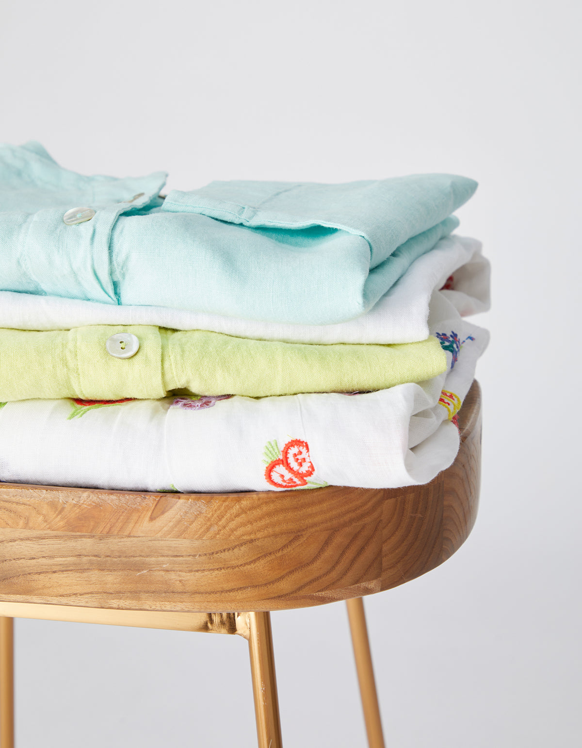 A image of a stack of folded clothes sits on a wooden stool. From top to bottom: light blue, white, lime green, and embroidered white shirts.