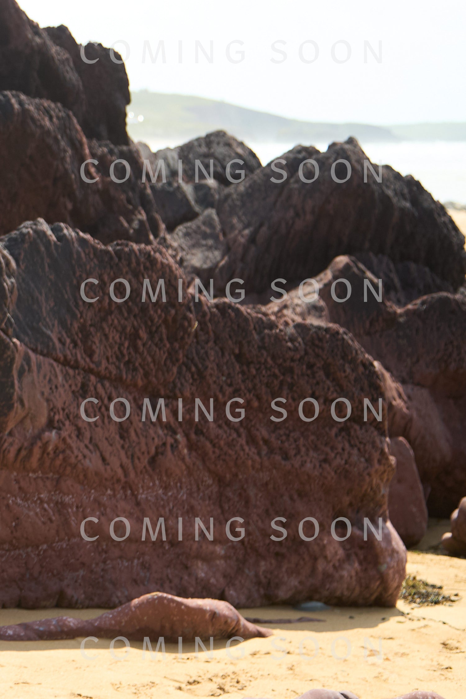 Large reddish-brown rocks on a sandy beach with greenery and ocean in the background evoke the artistry of Hand Crocheted Teardrop Earrings. The text "COMING SOON" is repeatedly overlaid on the image.