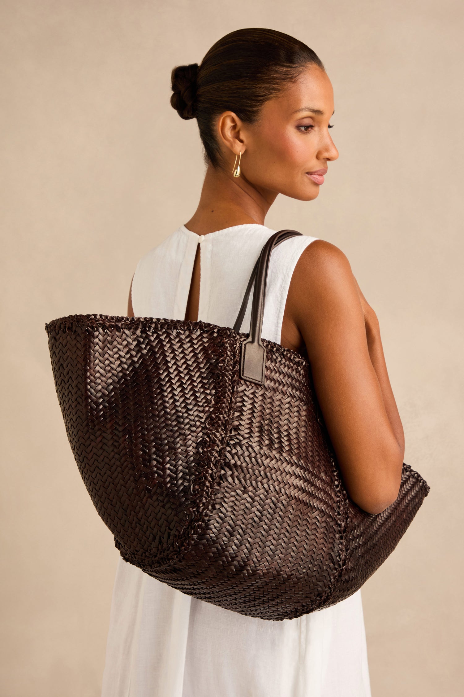 A woman in a white sleeveless dress holds the Teresa Woven Leather Bag—a large, dark brown piece that highlights exquisite handwoven craftsmanship—over her shoulder as she poses against a neutral background.