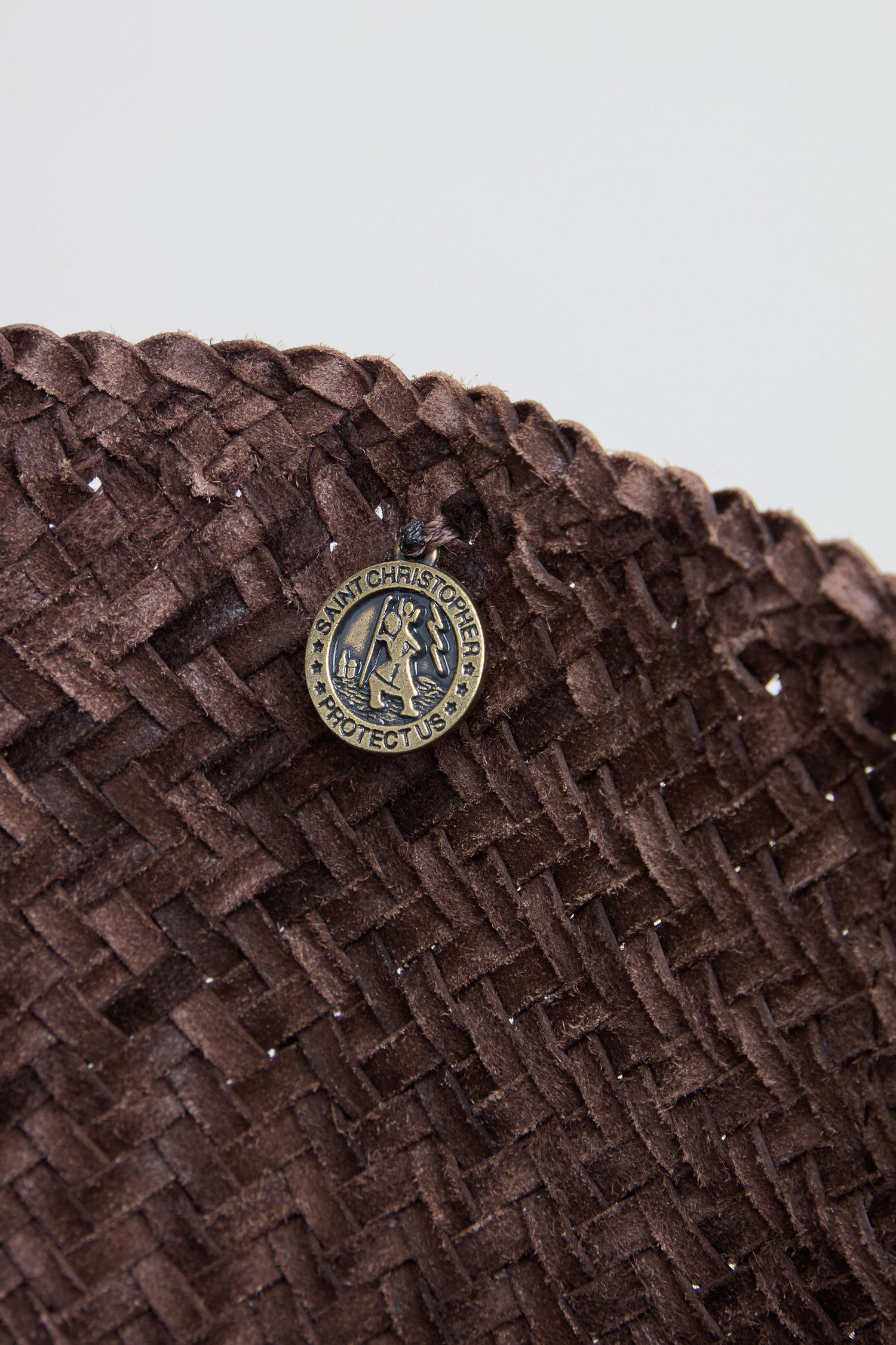 A close-up of the Teresa Woven Leather Bag highlights its handwoven craftsmanship, with a round Saint Christopher "Protector" pin attached near the top edge.