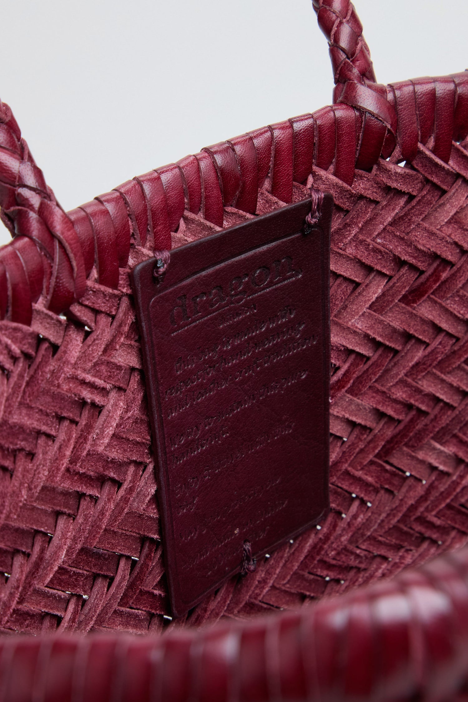 Close-up of a woven maroon handbag, identified as the Small Triple Jump Woven Leather Bag, with a rectangular leather tag attached to the interior featuring embossed text.
