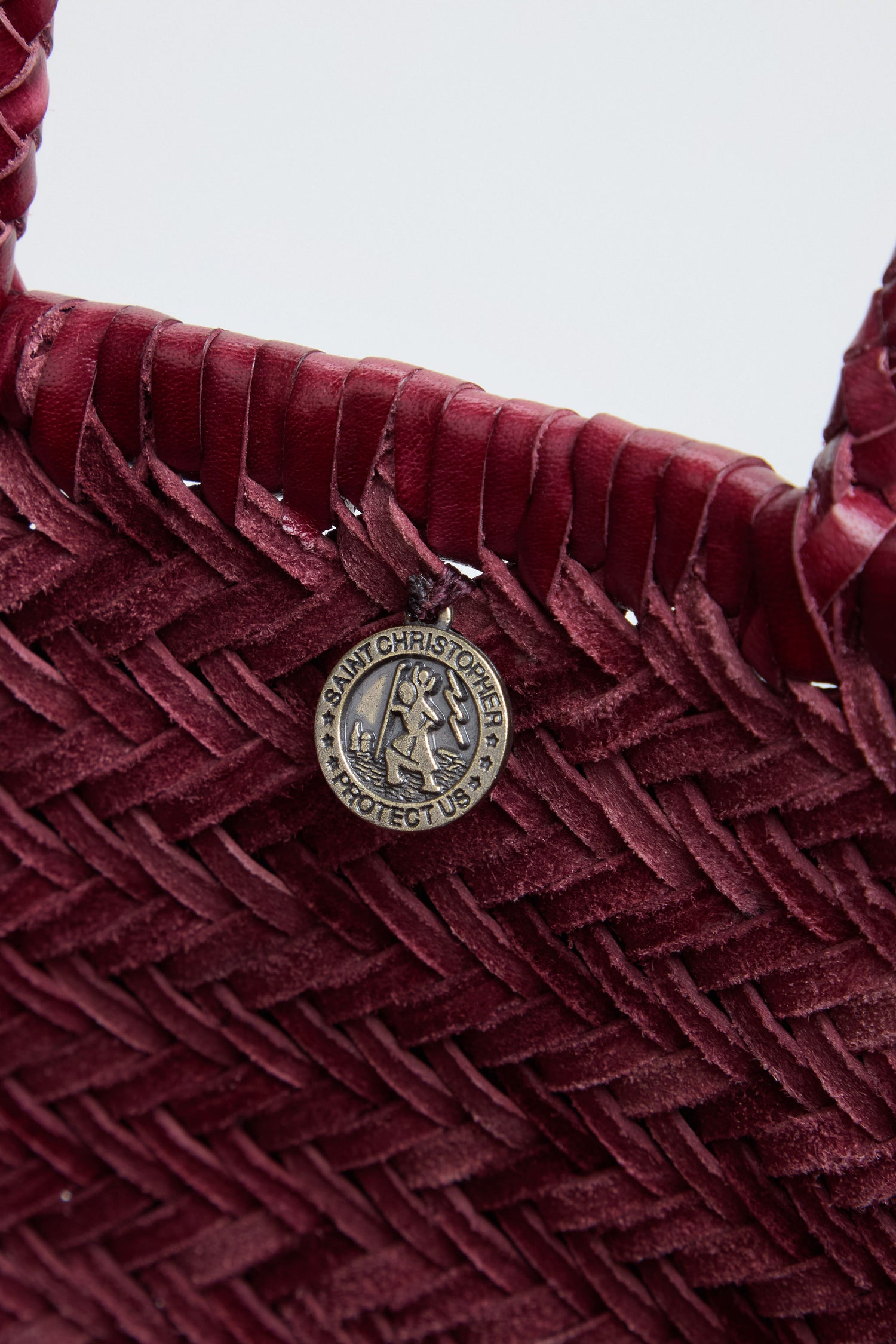 Close-up of a maroon Small Triple Jump Woven Leather Bag by Dragon Diffusion with a round silver Saint Christopher medal attached, featuring a detailed handwoven pattern and visible stitching.