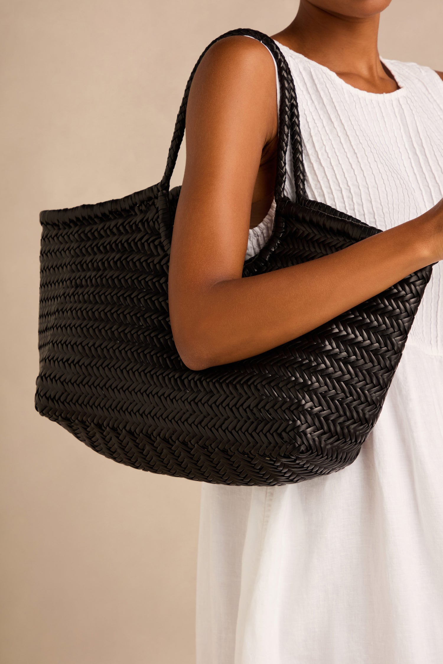 A person in a sleeveless white dress holds the Nantucket Woven Black Leather Bag over their shoulder against a neutral background.