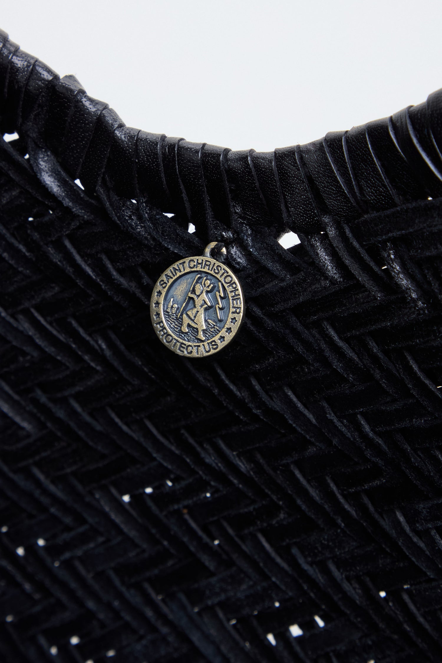 Close-up of the Nantucket Woven Black Leather Bag, its handwoven leather surface elegantly displaying a round metallic pin engraved with "Saint Christopher Protect Us" attached near the top edge.