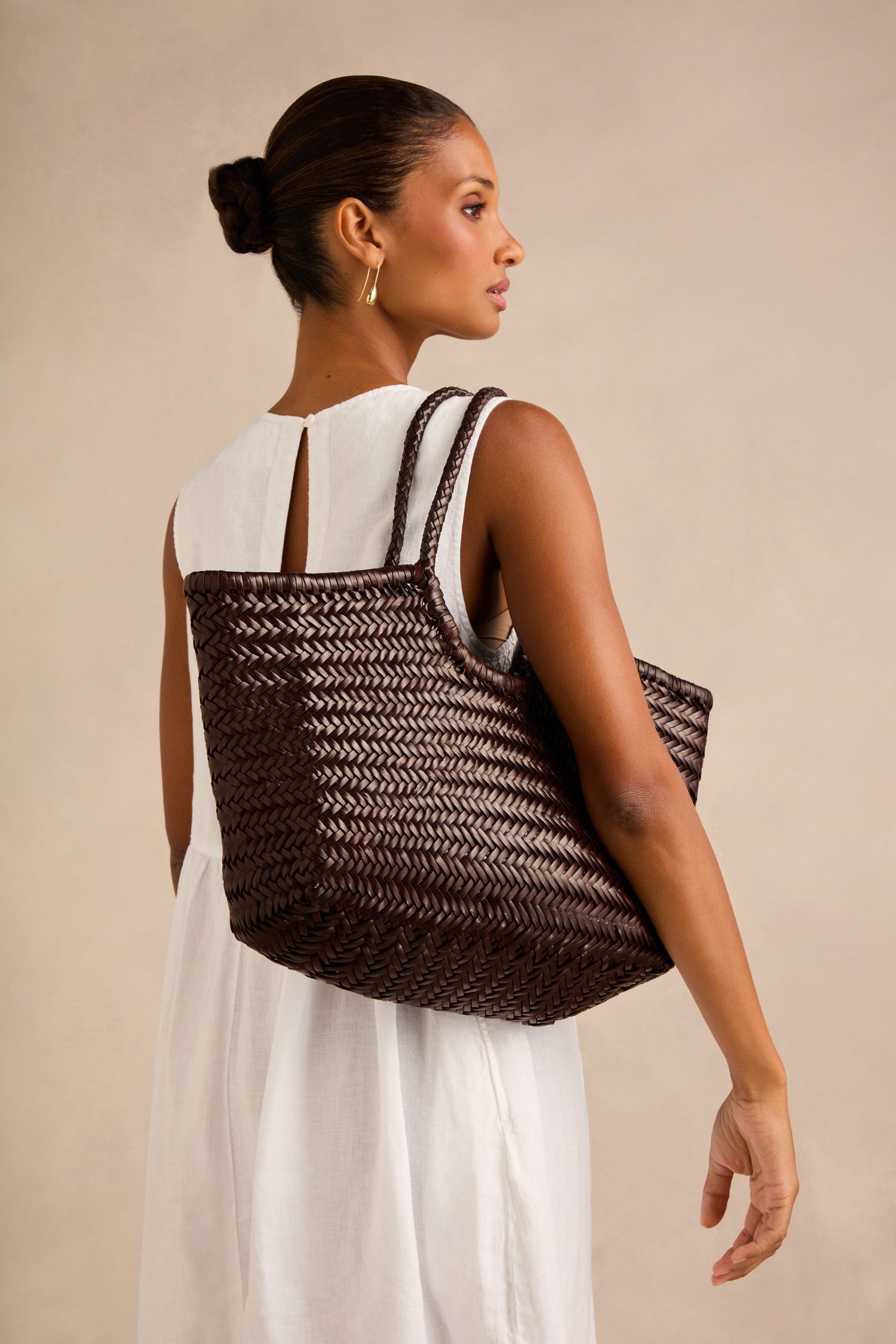 A woman in a white sleeveless dress carries the Nantucket Woven Dark Brown Leather Bag over her shoulder—its handwoven design echoing the timeless elegance of Dragon Diffusion—as she stands against a plain beige background.