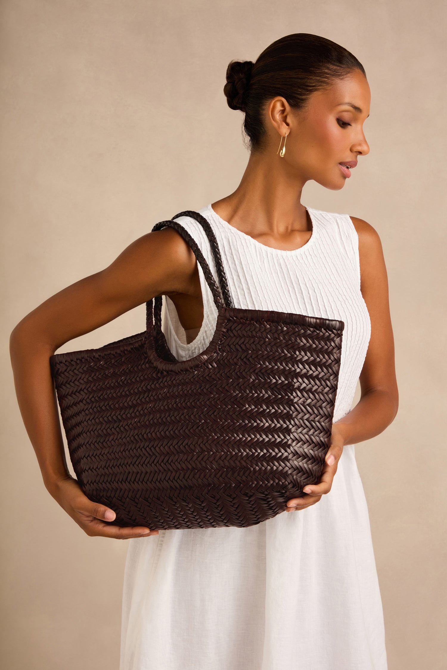 Woman in a white sleeveless dress holds a large, dark brown Nantucket Woven Dark Brown Leather Bag against a plain beige background.