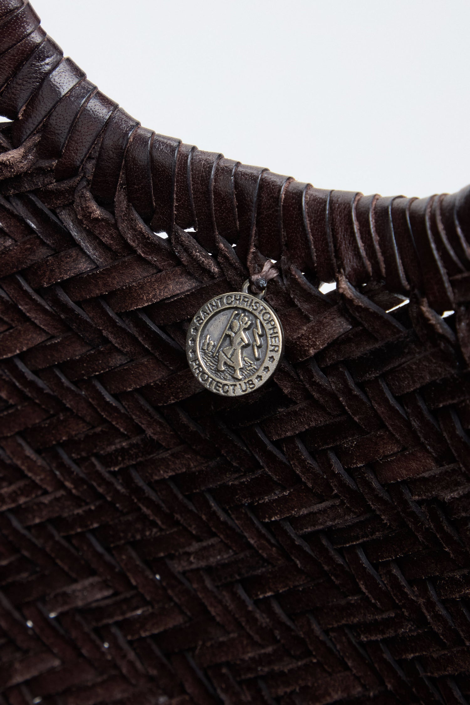 Close-up of a woven dark brown leather strap from the Nantucket Woven Dark Brown Leather Bag, featuring a round metallic Saint Christopher medallion attached.