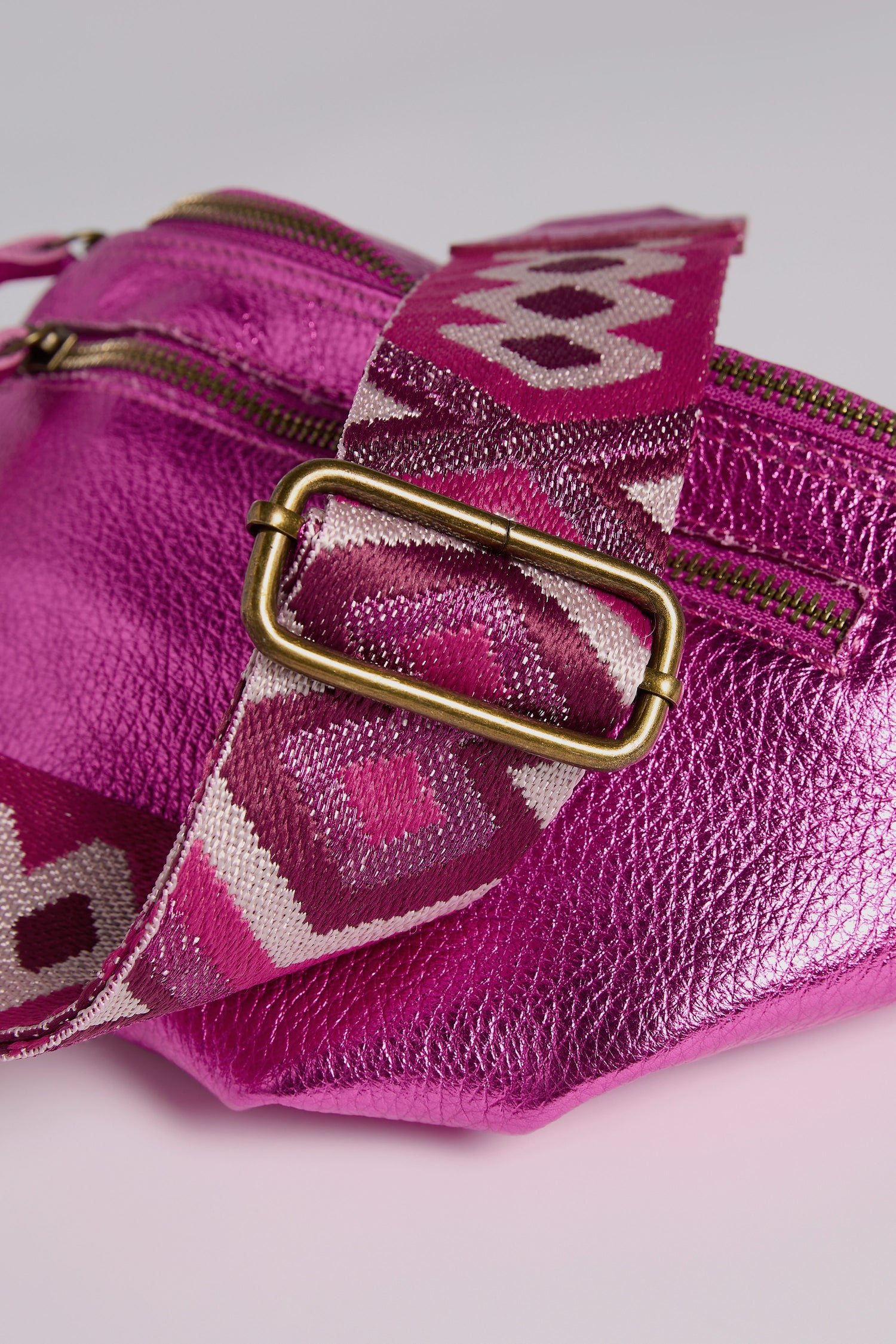 Check out the Doris Maroquinerie's Metallic Leather Crossbody Bag. This pink metallic bag features a patterned fabric strap, brass buckle, and visible zipper, topped with an iridescent leather finish for an elegant yet modern touch.