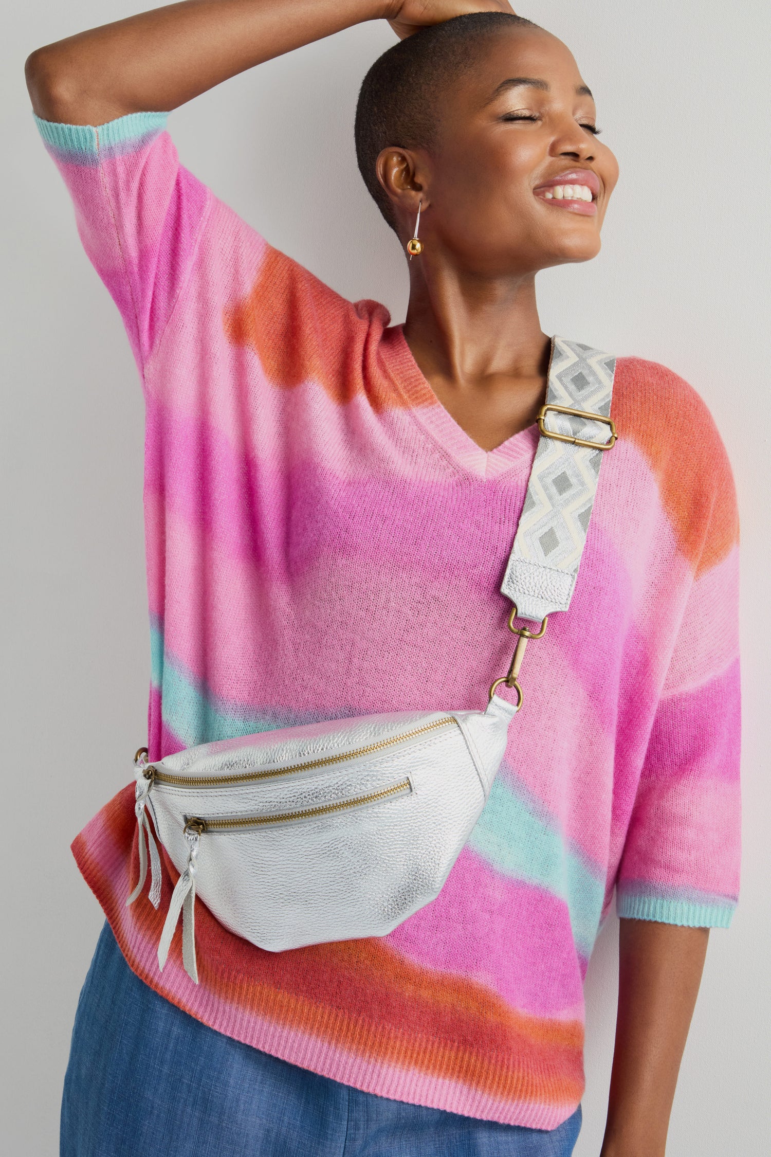 Person wearing a colorful striped sweater and blue pants poses with a Metallic Leather Crossbody Bag from Doris Maroquinerie.