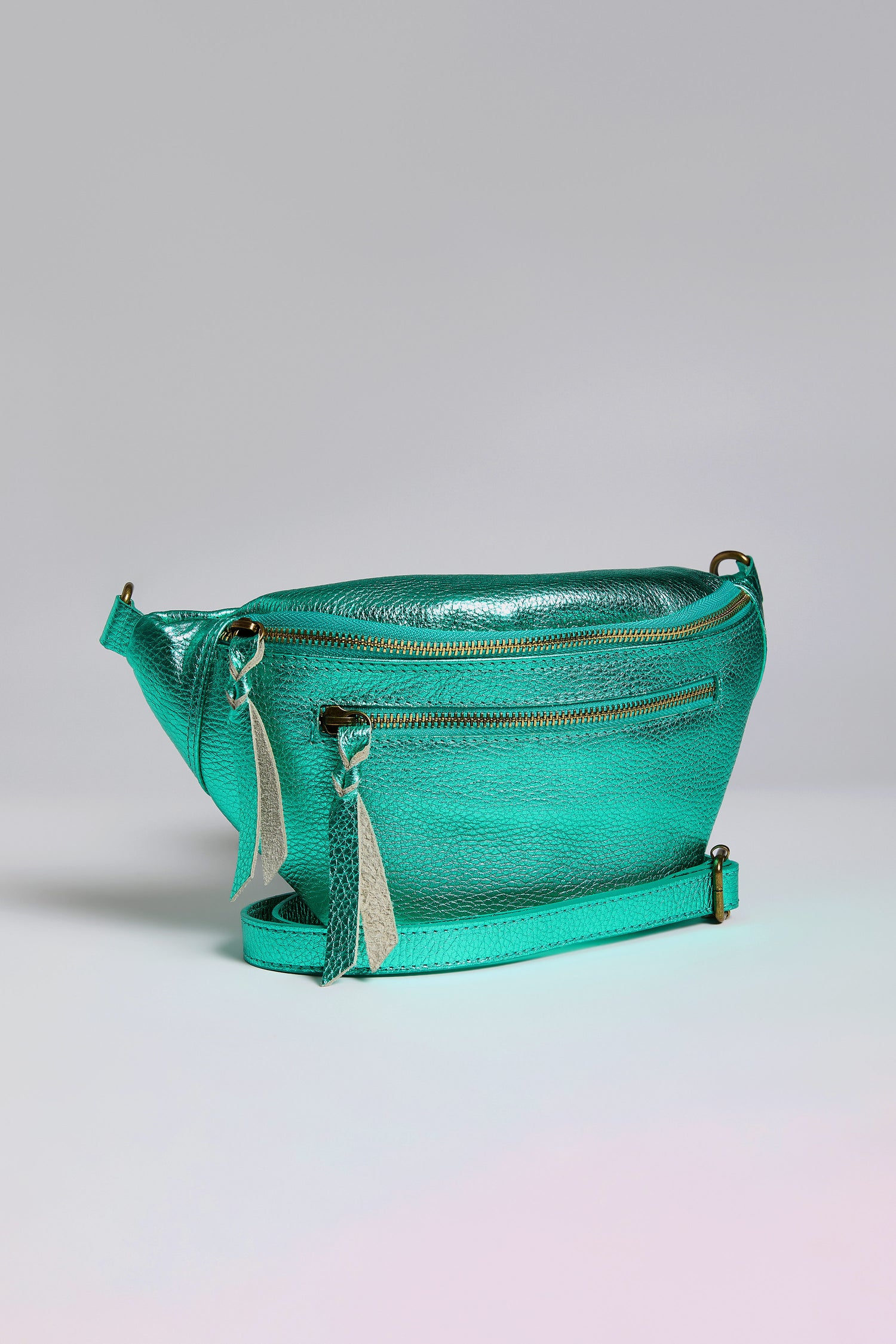 The Metallic Leather Crossbody Bag is crafted from iridescent teal leather, featuring a front zipper pocket and two decorative tassels. It stands elegantly against a plain gray background, embodying both style and functionality.