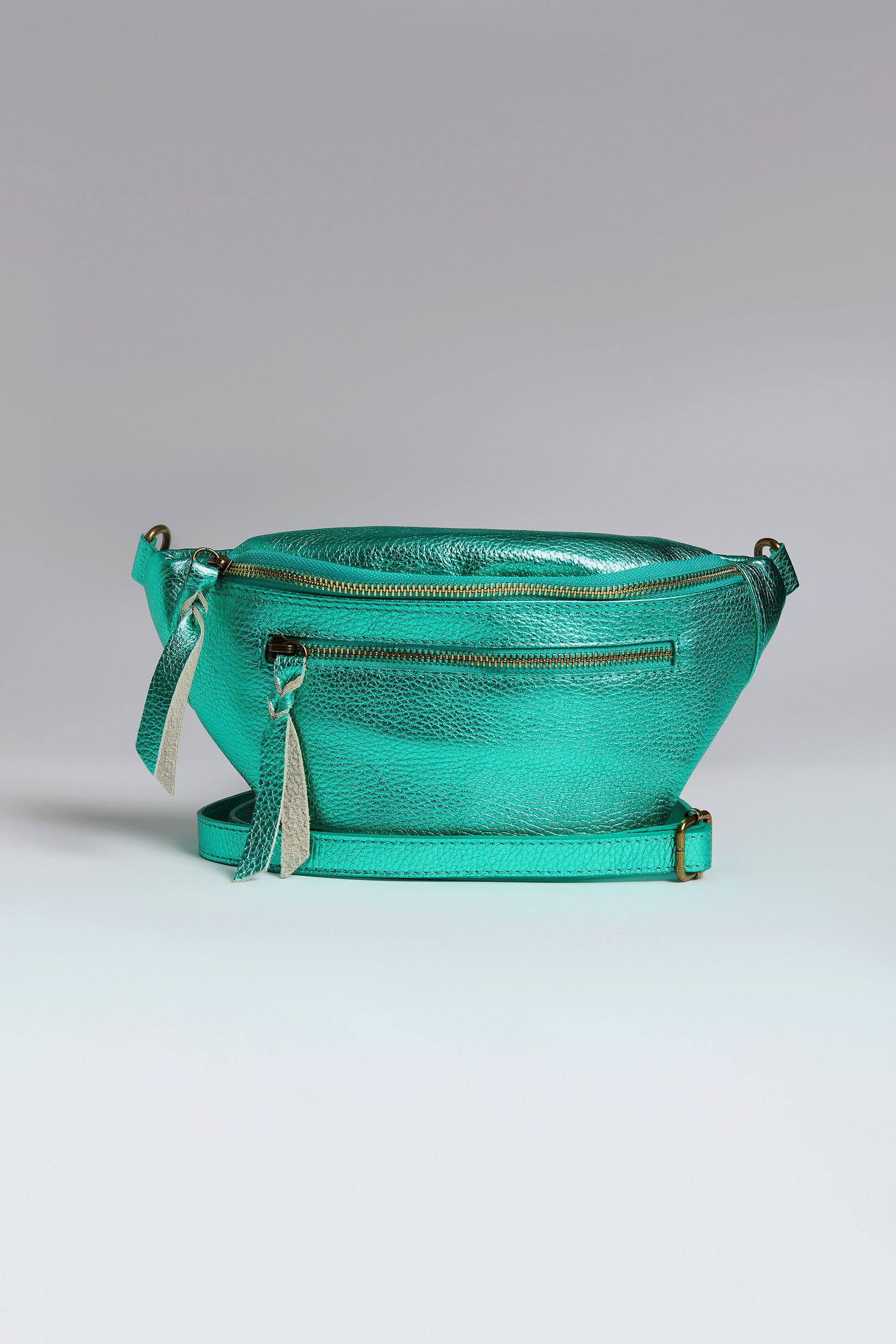 The Metallic Leather Crossbody Bag in metallic teal features iridescent leather, a front zipper pocket, an adjustable strap, and two tassel zipper pulls against a neutral background.