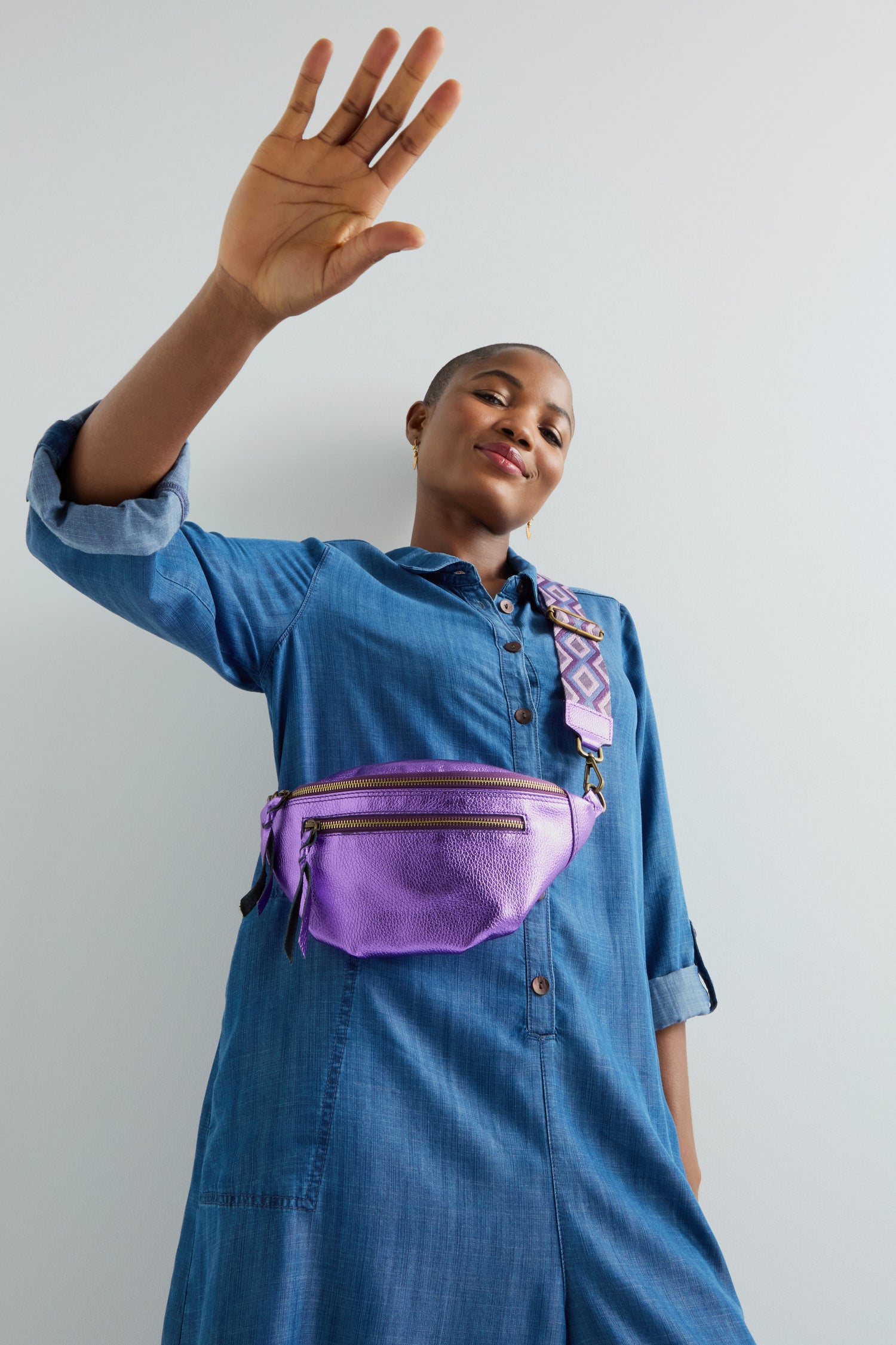 A person in a denim dress raises a hand, displaying their metallic leather crossbody bag with a patterned strap from Doris Maroquinerie.