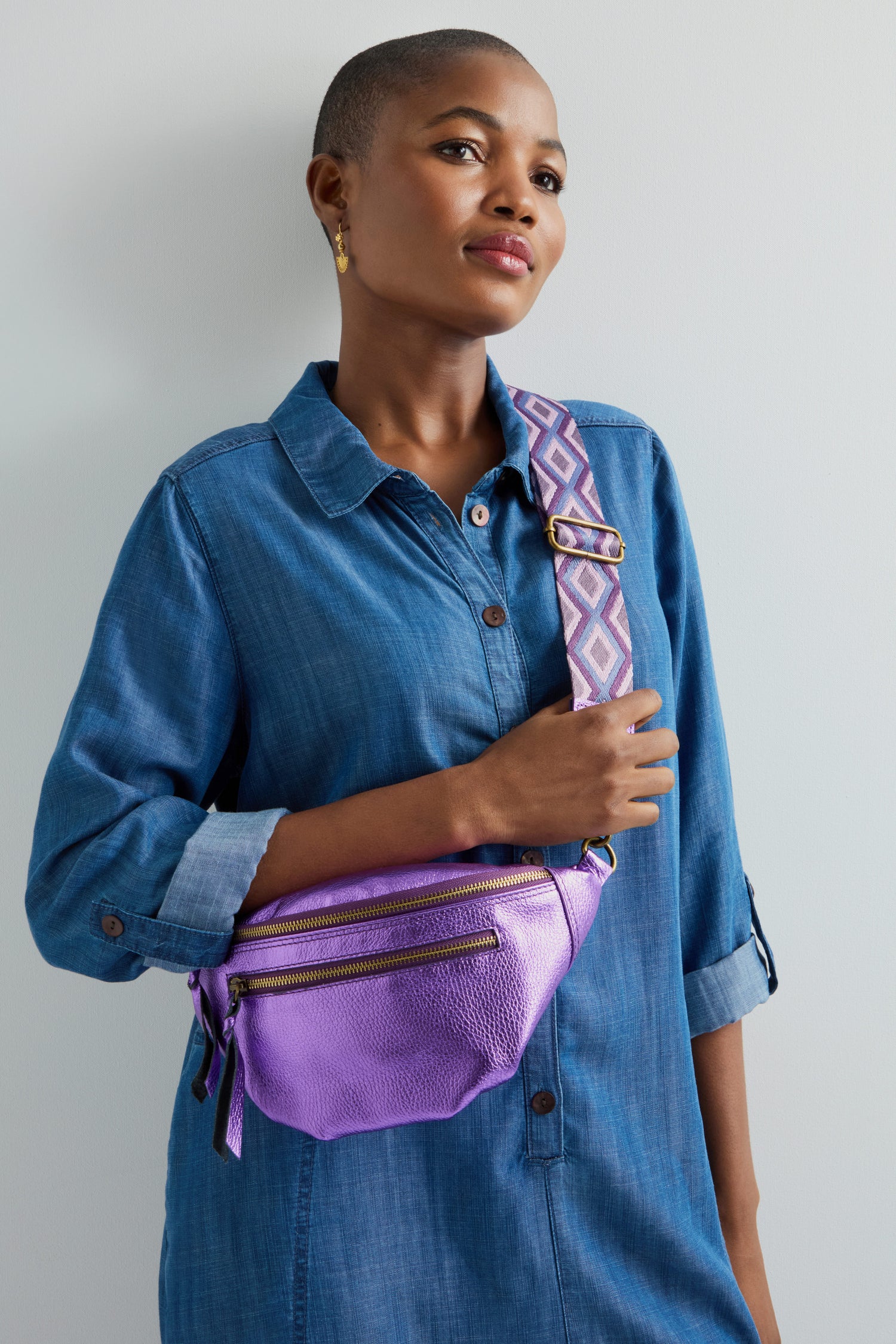 A person in a blue denim dress carries the stylish Metallic Leather Crossbody Bag with a patterned strap, enhancing their elegant look.