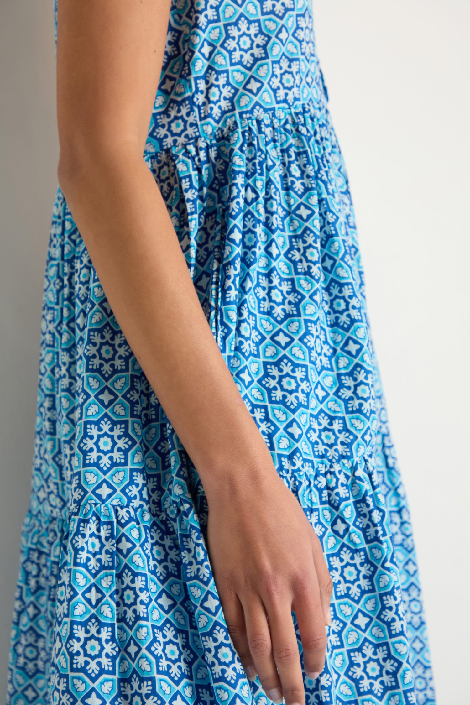 A person models the Floral Tiered Dress, a sleeveless blue and white outfit, standing against a plain white background with their arm resting by their side.