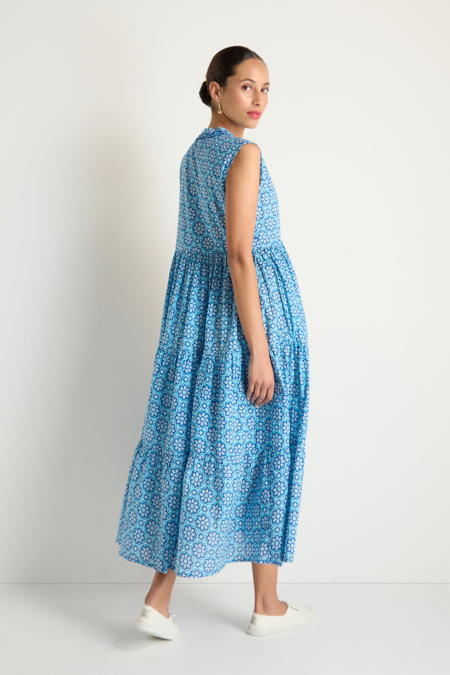 A woman models the Floral Tiered Dress, a sleeveless blue and white floral piece, paired with white sneakers as she stands against a plain white background, glancing over her shoulder.