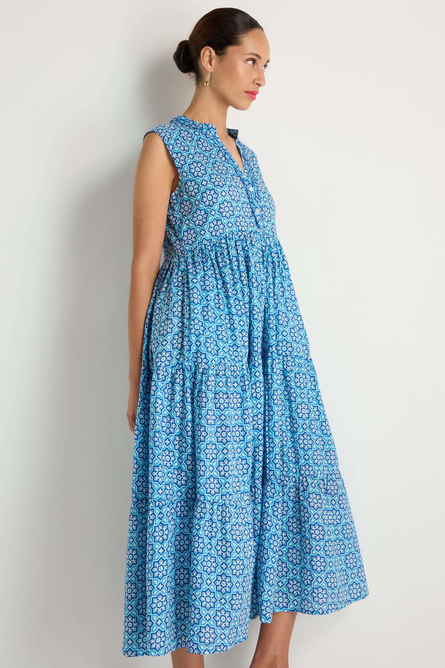 A woman stands against a white wall wearing the Floral Tiered Dress, featuring blue and white florals, a v-neckline, gathered waist, and sleeveless tiered maxi silhouette.