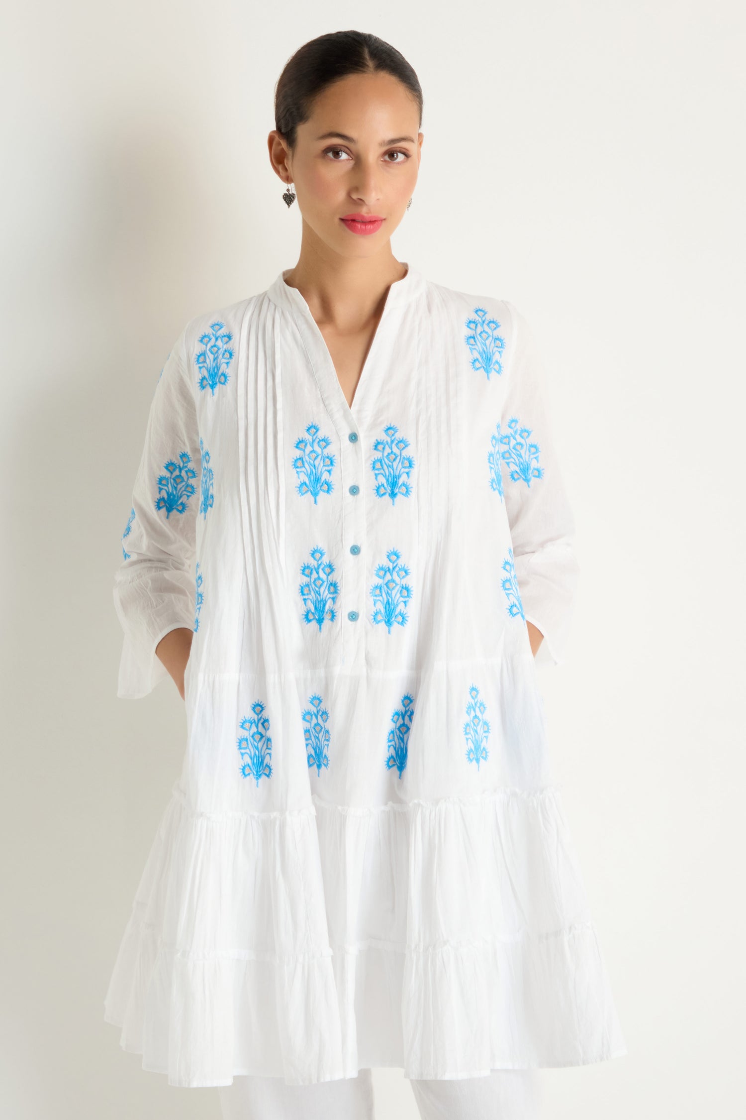 A woman stands against a white background wearing the Embroidered Floral Flared Sleeve Tunic, featuring pure cotton fabric and delicate blue floral designs.