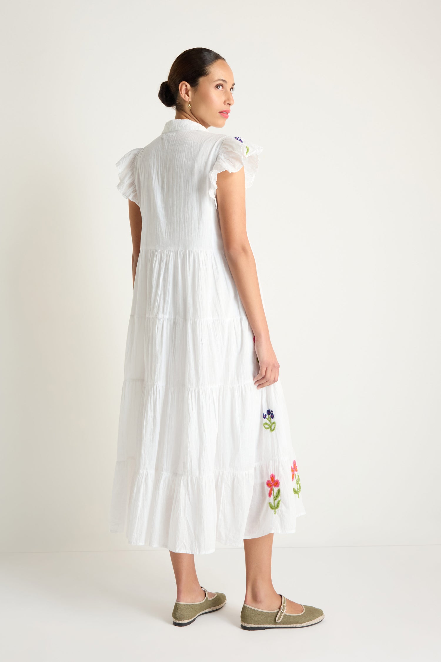A woman stands in a minimalist setting, looking over her shoulder in the Embroidered Flower Frill Sleeve Dress featuring a white tiered skirt and delicate floral embroidery.