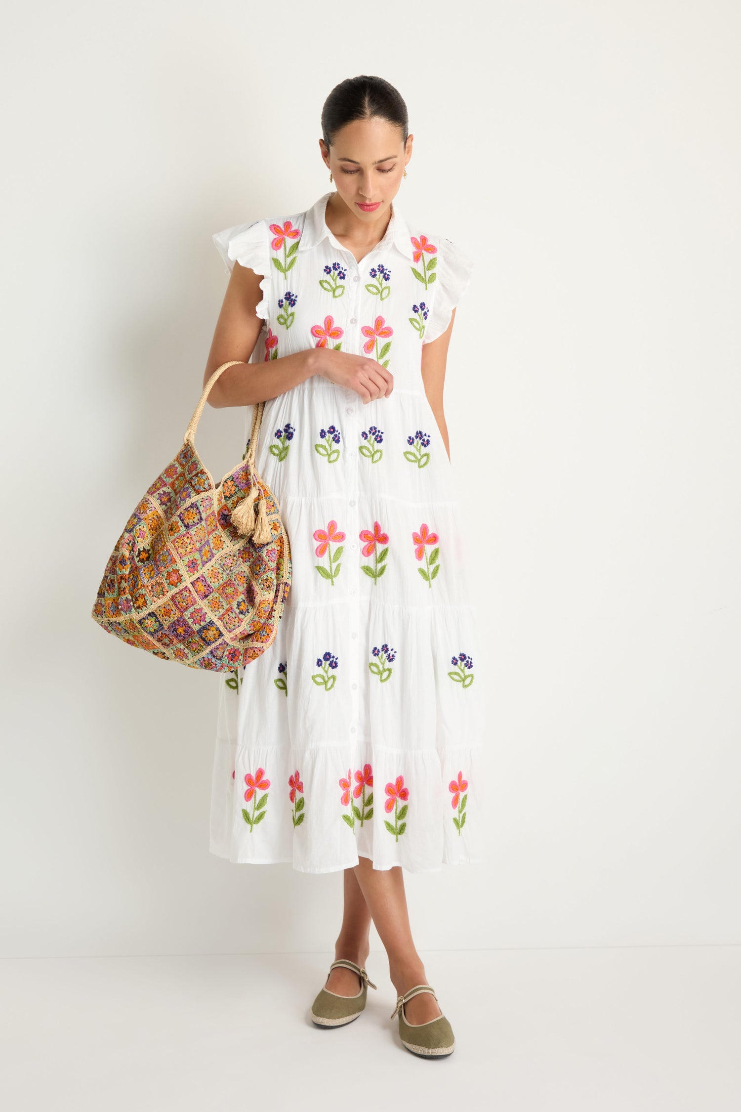 A woman stands against a white wall wearing the Embroidered Flower Frill Sleeve Dress, featuring colorful floral details, and holds a large patterned shoulder bag with beige flat shoes.
