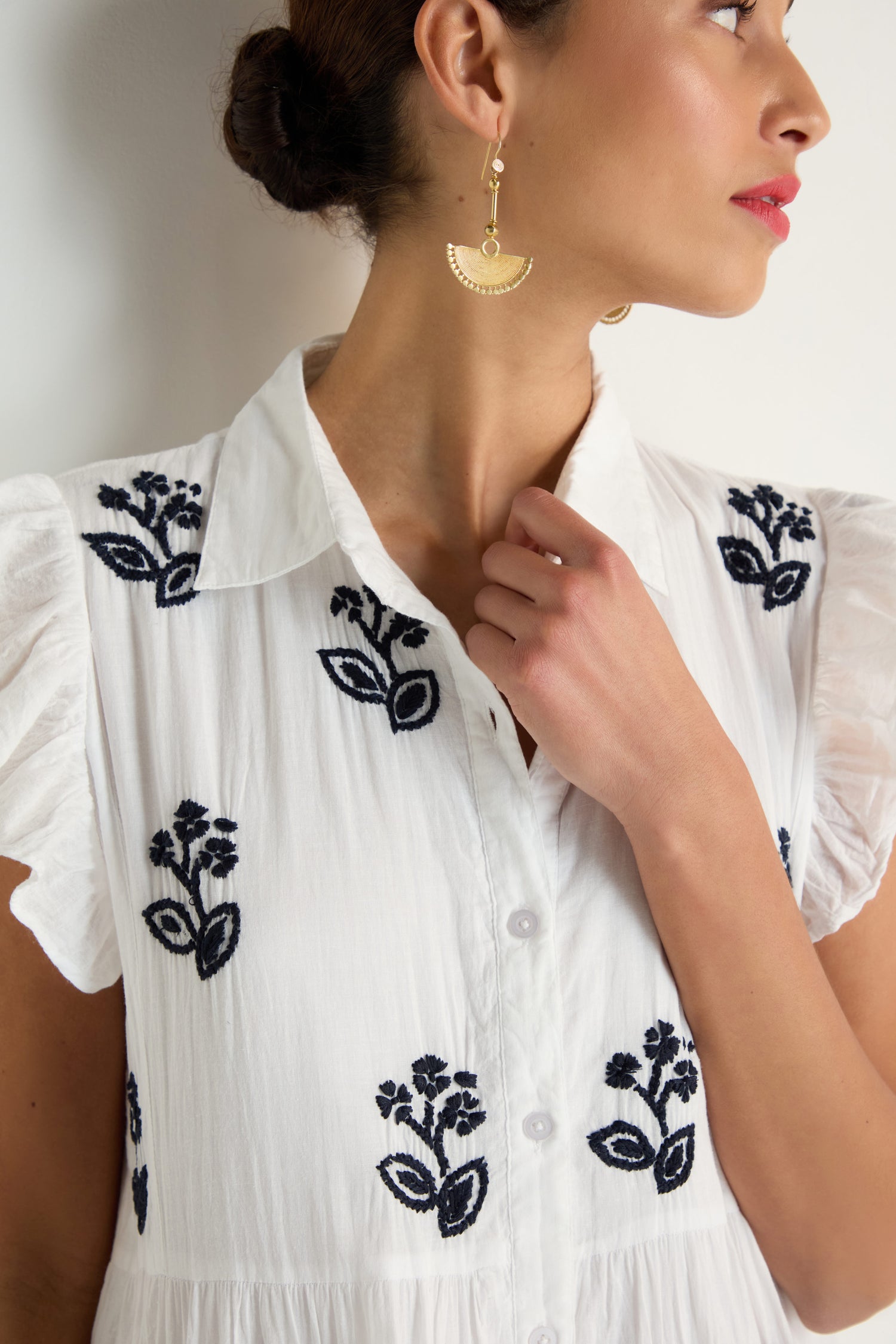 Woman wearing the Embroidered Navy Floral Frill Sleeve Dress, styled with gold half-moon earrings and her hair tied back, gazing to the side.
