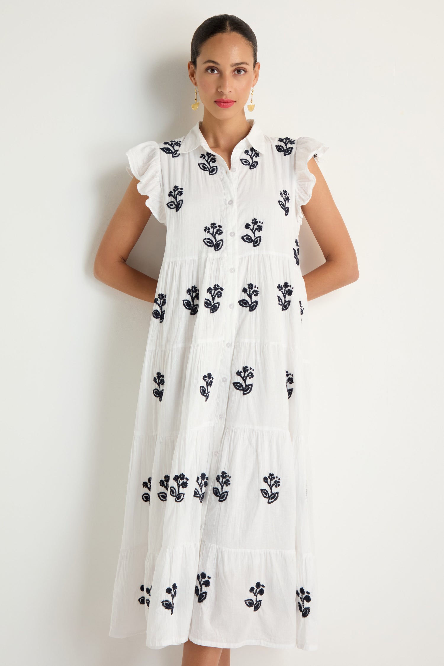 A woman stands against a white wall wearing the Embroidered Navy Floral Frill Sleeve Dress—a button-down, sleeveless cotton midi with navy floral embroidery and ruffled shoulders.