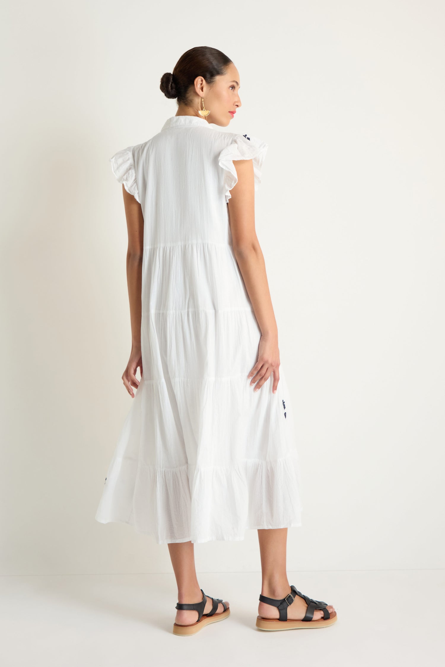 Woman standing in an Embroidered Navy Floral Frill Sleeve Dress, black sandals, and gold earrings, facing away from the camera against a plain white background.