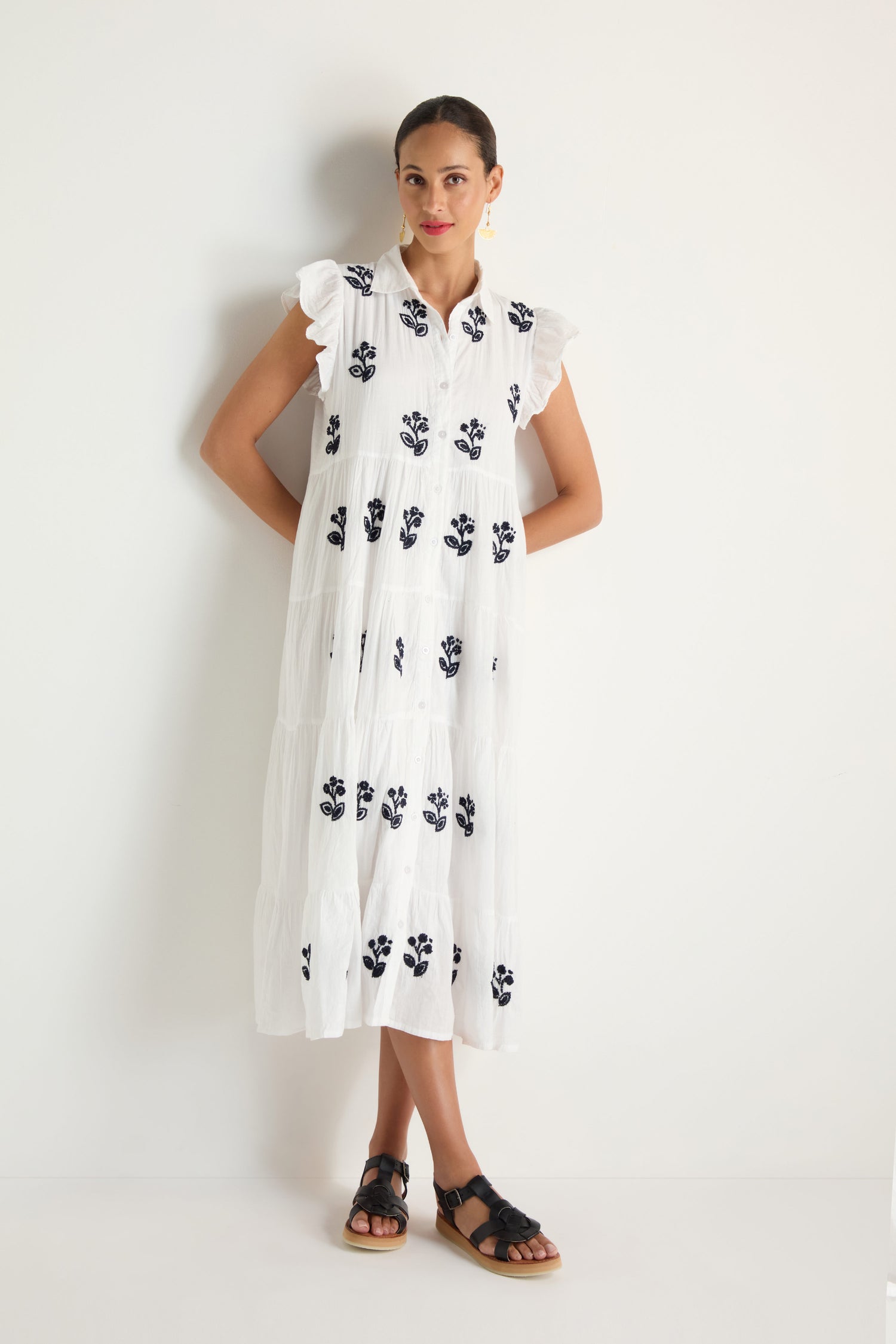 A woman stands against a white wall wearing the Embroidered Navy Floral Frill Sleeve Dress paired with black sandals.