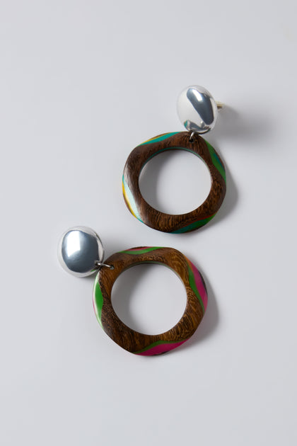 The Rainbow Spheres Clip-On Earrings feature round silver studs attached to oversized wooden hoops with irregular circular shapes and green and pink accents, shown on a plain white background.