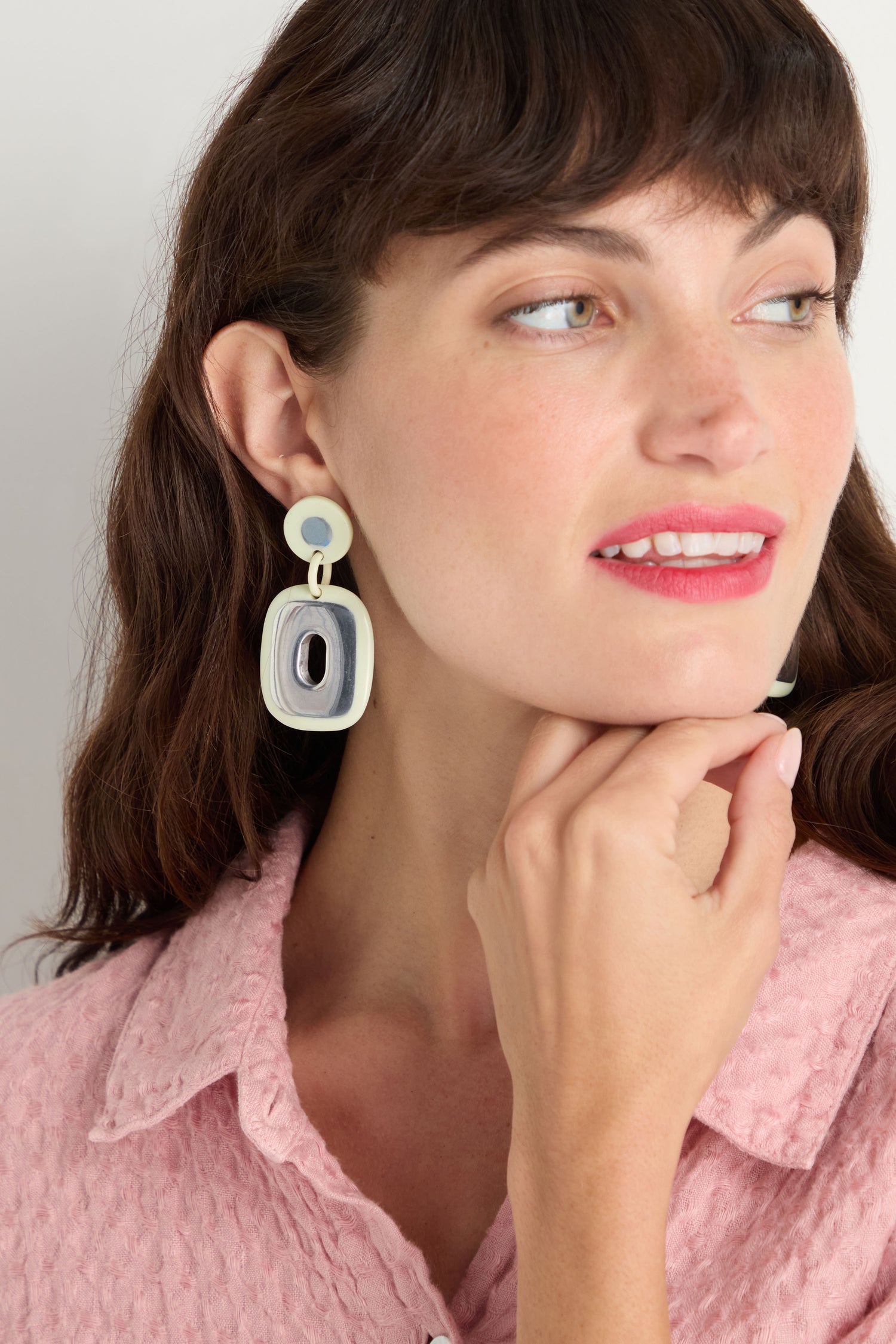 A woman wears Deco Earrings, featuring large recycled aluminium pieces, paired with a textured pink shirt. She looks right, touching her chin thoughtfully.