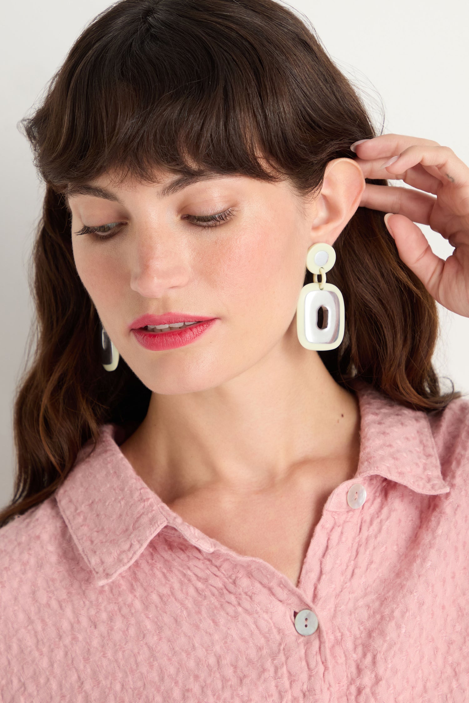 Woman with wavy brown hair wears a pink textured button-up shirt and Deco Earrings made from recycled aluminium, looking down and touching one statement earring.