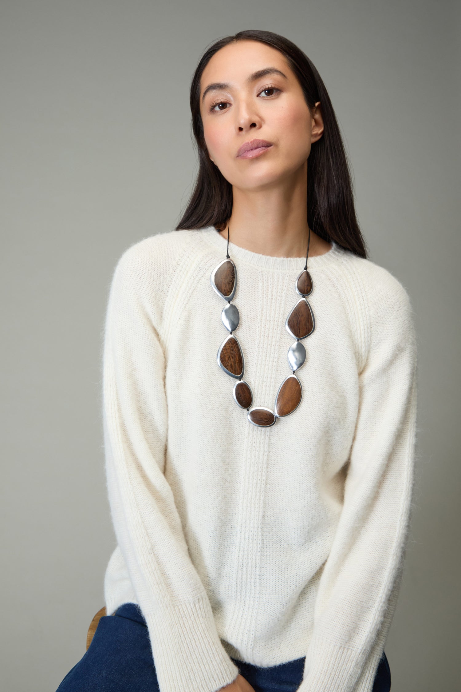A woman with long dark hair, in a cream sweater and blue jeans, wears the Wooden Fusion Pebble Necklace by Ernesto de Barcelona—featuring brown and silver beads—as she sits against a neutral background.