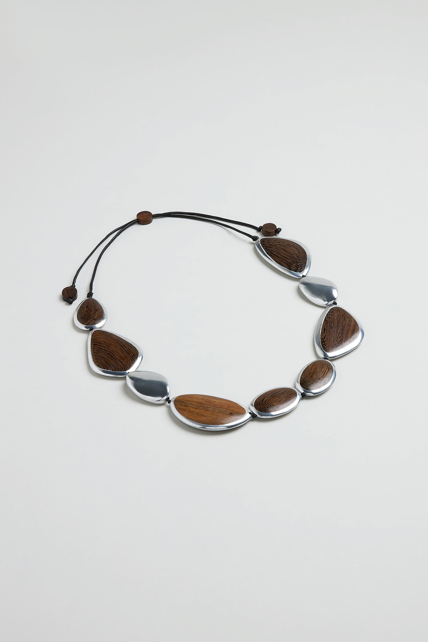 The Wooden Fusion Pebble Necklace showcases alternating oval and teardrop-shaped brown wooden and silver metal accents on a black adjustable cord, offering sculptural style inspired by Ernesto de Barcelona.