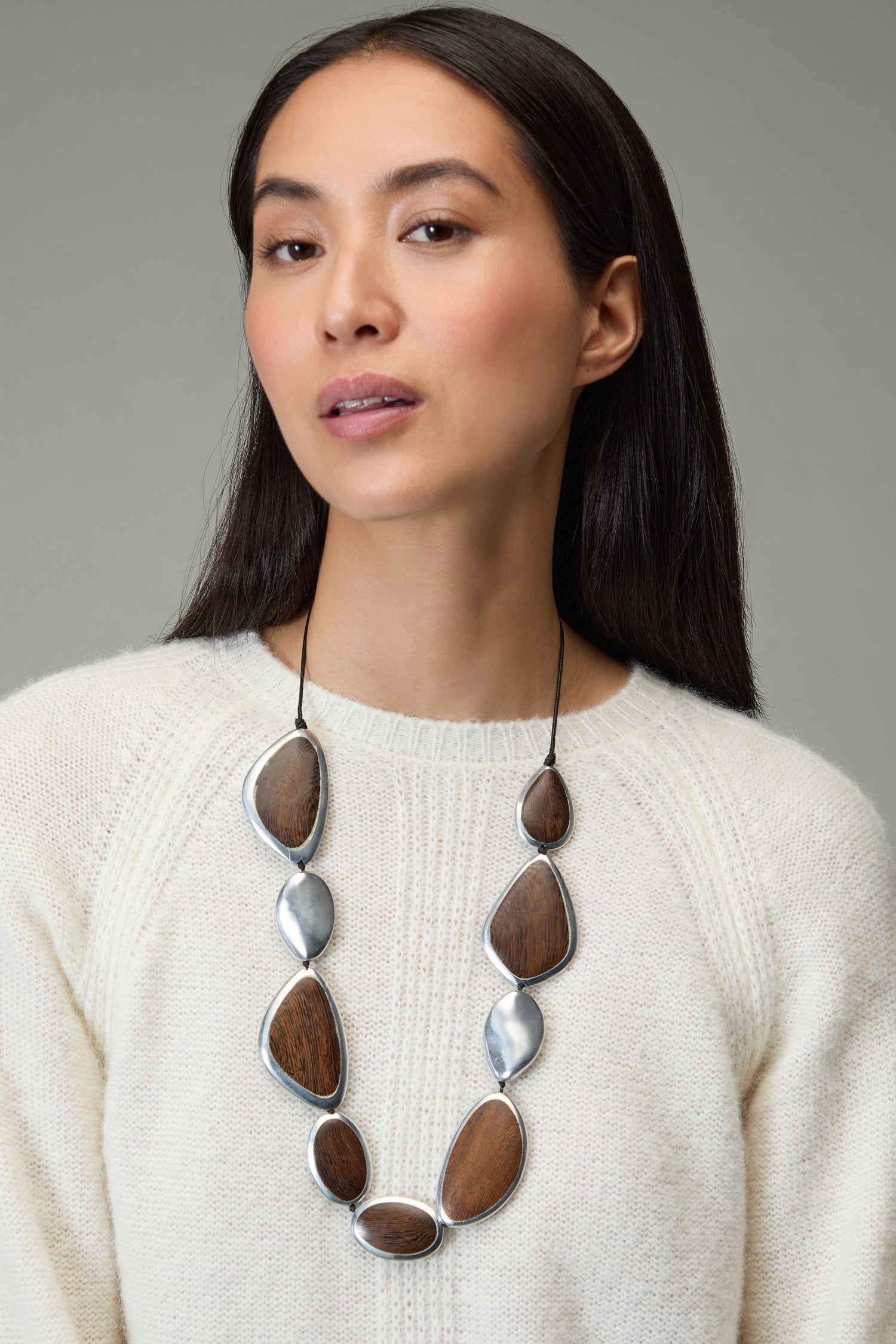 A woman with long dark hair wears a cream sweater and the Wooden Fusion Pebble Necklace, featuring large, irregular wooden and silver pieces.