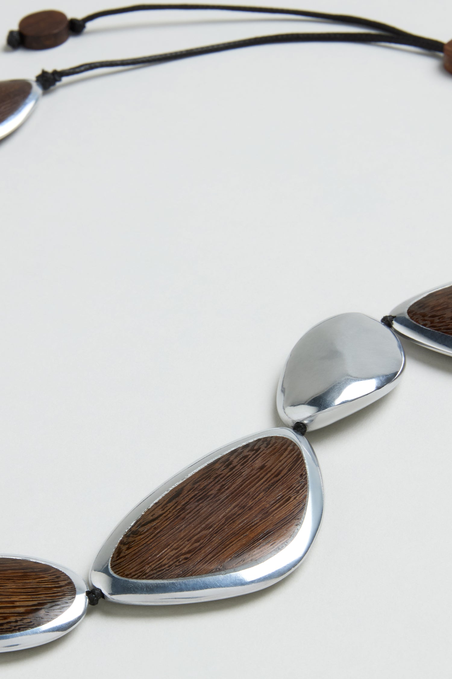 Close-up of the Wooden Fusion Pebble Necklace, featuring teardrop-shaped metal and wood segments on a black cord, shown against a plain light background for a sculptural jewelry look.