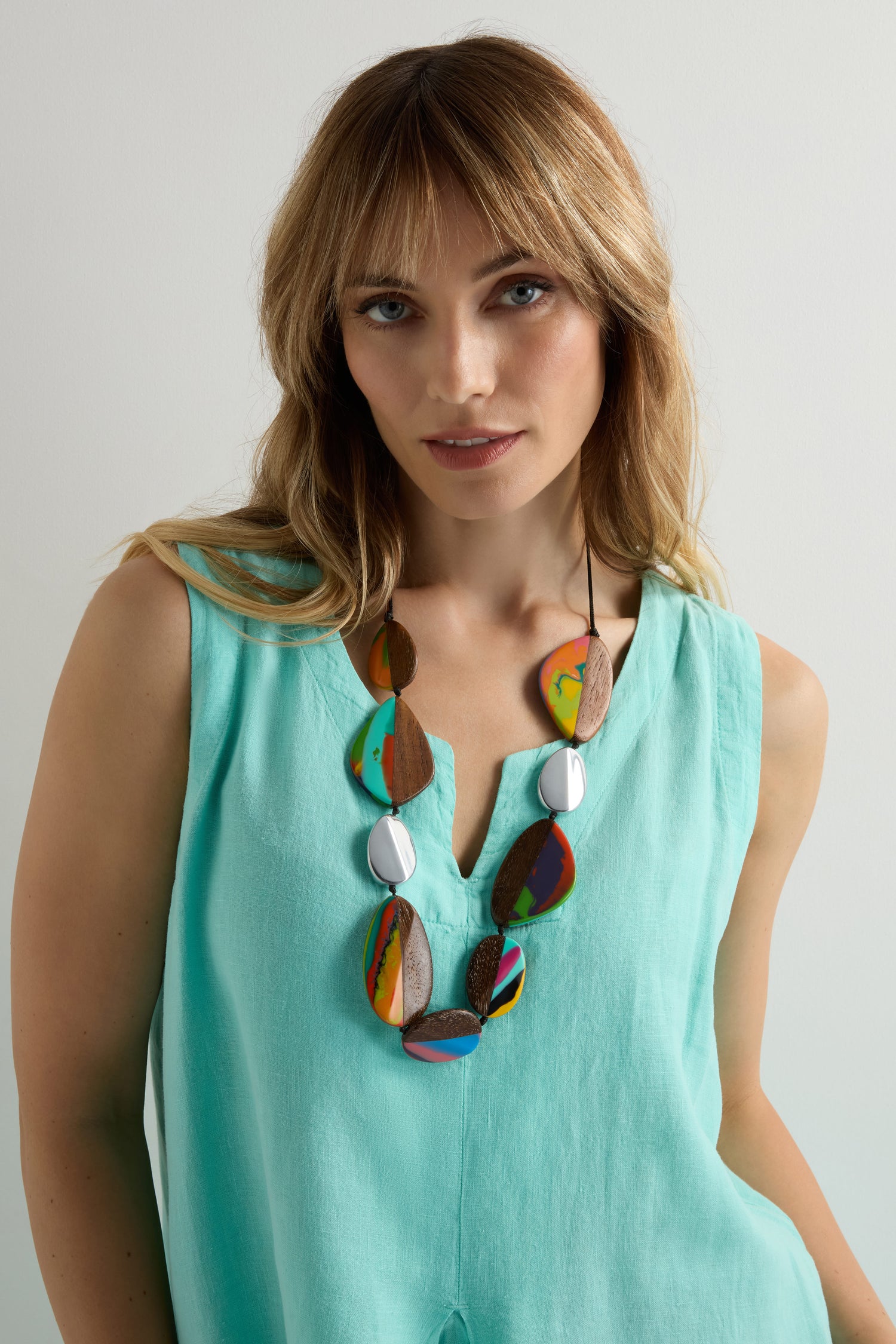 Woman in a sleeveless light turquoise top, wearing a Marble Resin Pebble Necklace by Ernesto de Barcelona, crafted from marbled resin, stands against a plain light background.