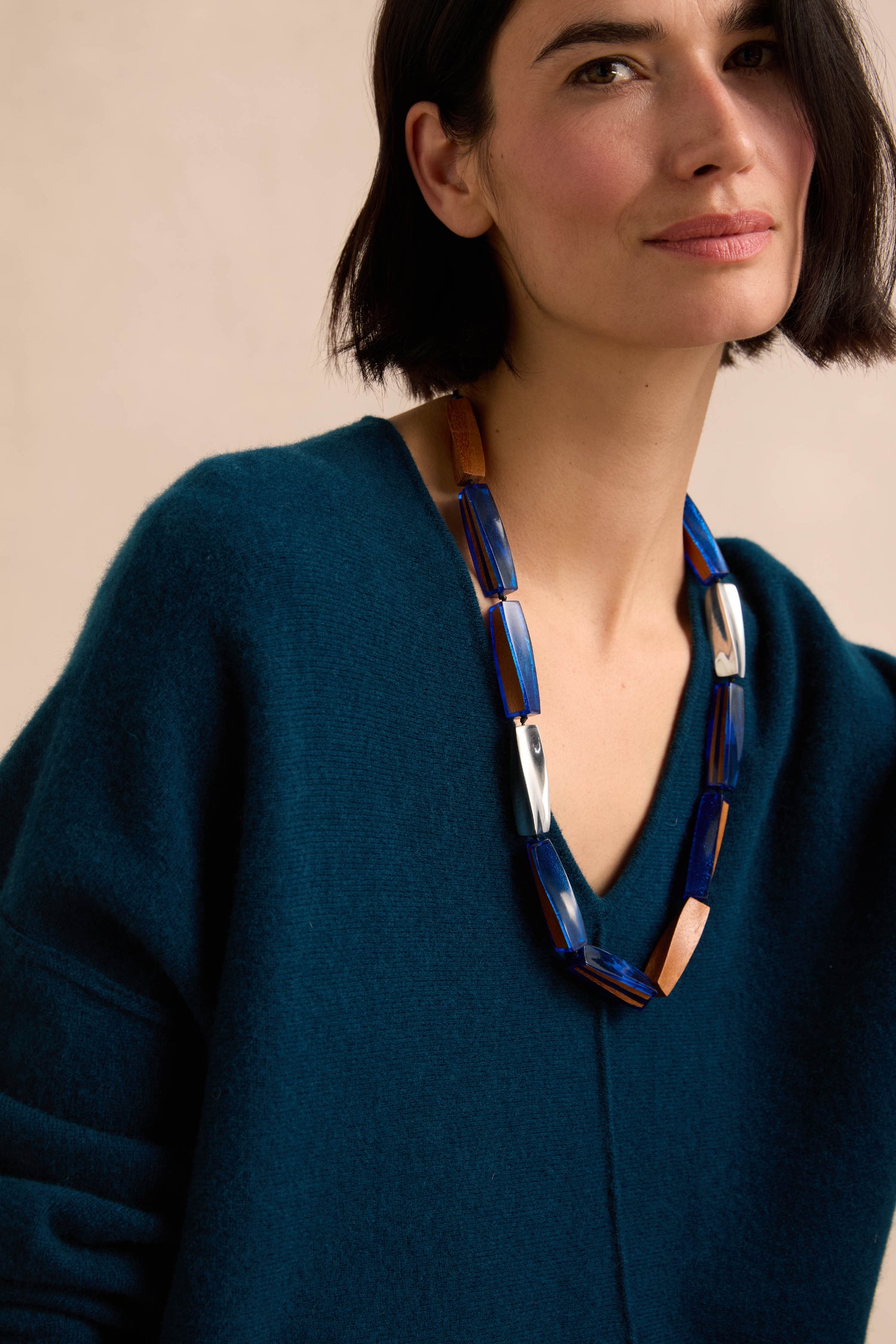 A woman with short dark hair wears a teal V-neck sweater and the Twisted Shapes Necklace—an elongated wood piece featuring chunky blue and brown beads—while looking at the camera.