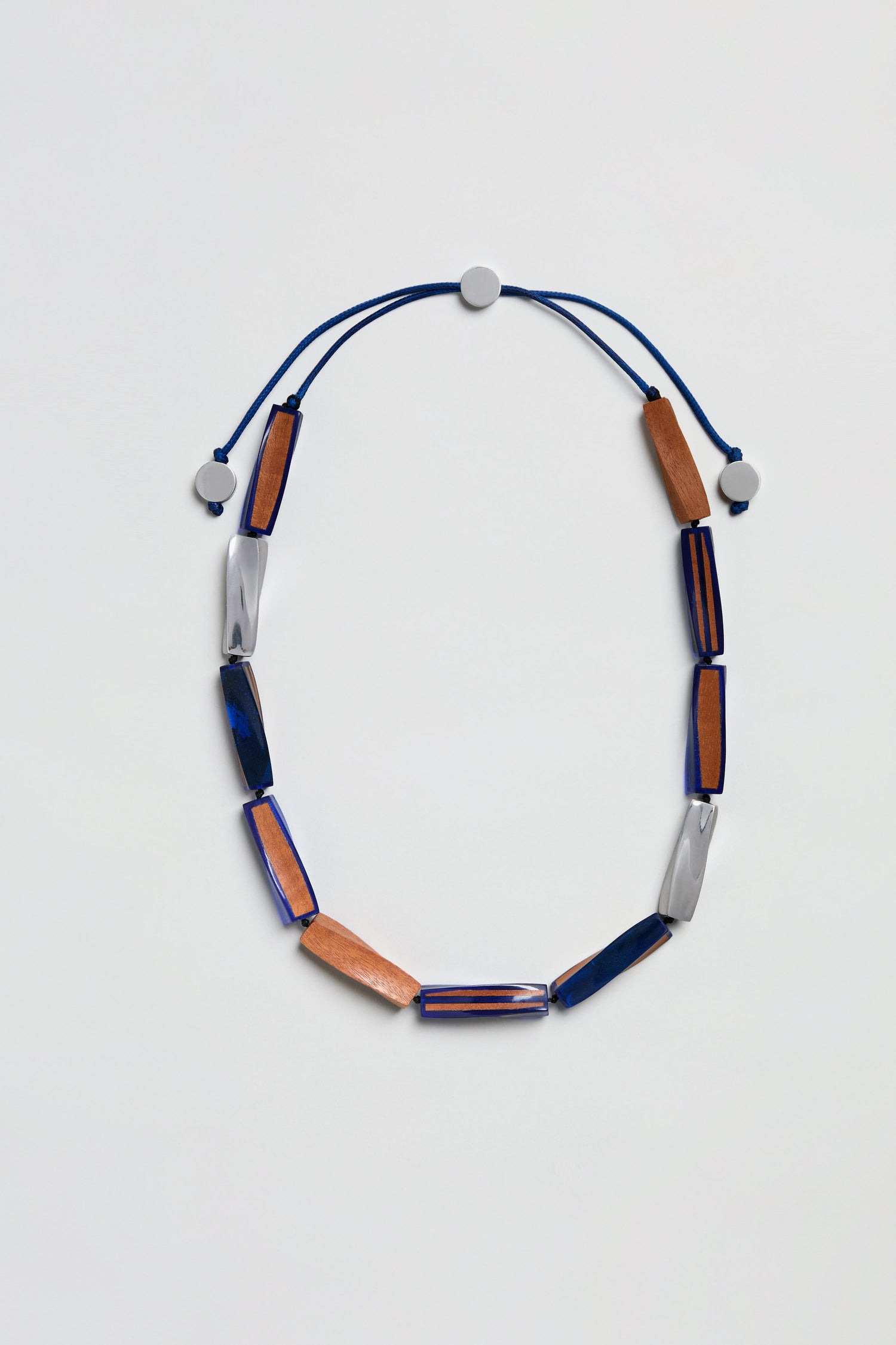 The Twisted Shapes Necklace is a stylish adjustable long necklace, featuring alternating rectangular brown, blue, and silver beads on a blue cord with two round adjustable sliders. This elongated wood necklace combines contemporary design with natural elements to create a unique finishing touch.