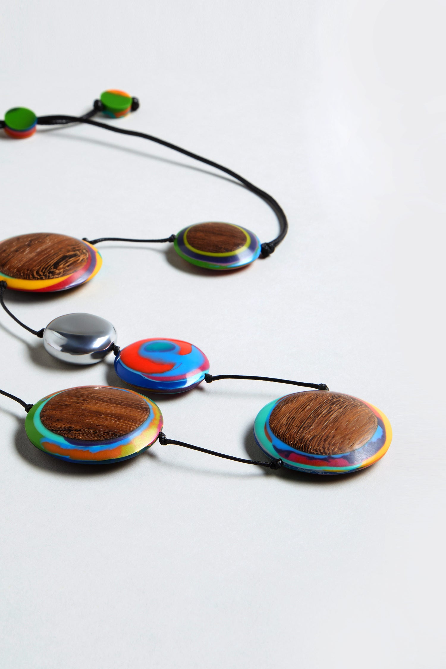 The Planetary Necklace features large, round beads with colorful patterns and wood textures, plus a metallic recycled aluminium disc, all on a black cord and displayed on a white surface.